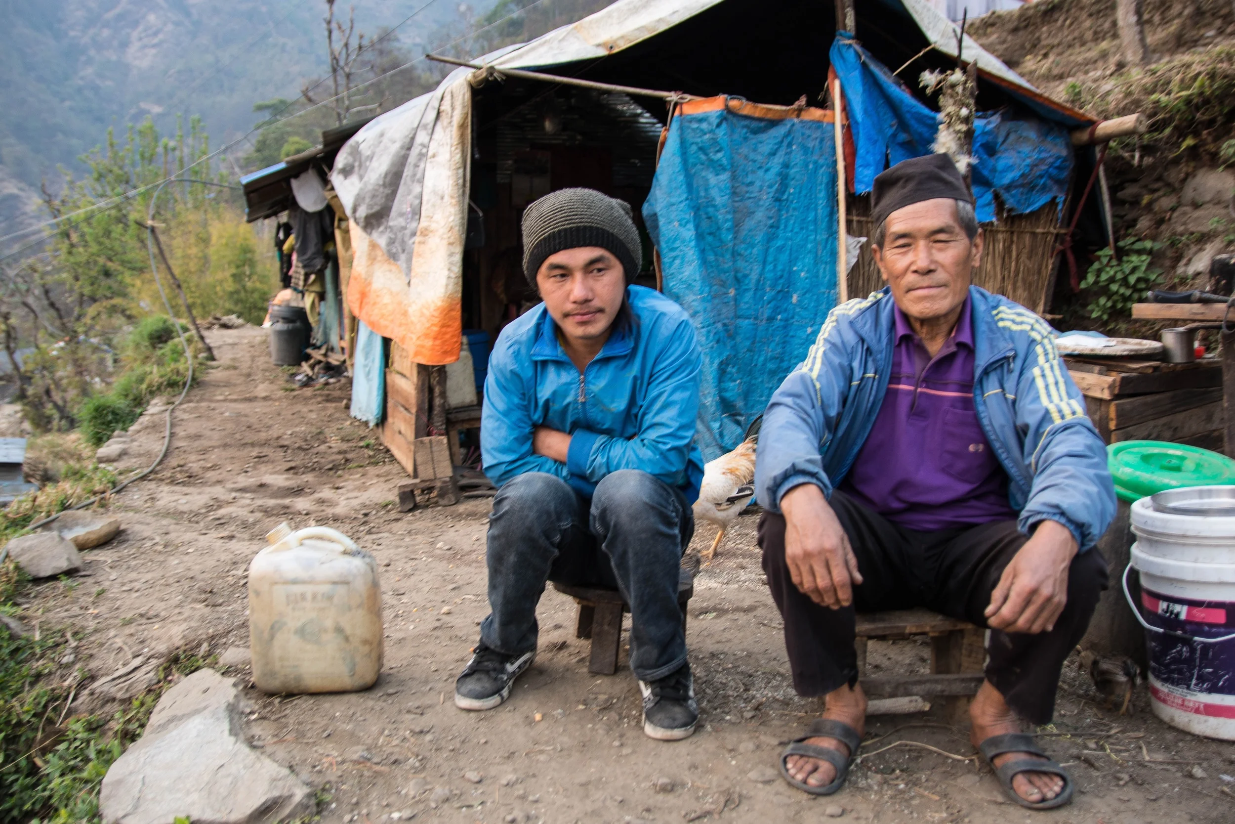 Pream Bahadur Tamang, right, and his family are still living under their tarpaulin-covered shelter. He has been watching his neighbors rebuild their houses and has decided to wait and see if their homes last through the upcoming monsoon season before he decides whether or not to rebuild his own house. His family, including his son Bir, left, disagree with his decision - Photo by Mark Morrison.