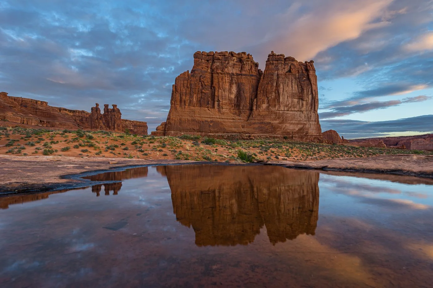 A wide landscape photograph capturing the massive sandstone Courthouse Towers in Arches National Park at sunrise, with pink-tinged clouds in a blue sky perfectly mirrored in a large, clear water pothole in the foreground.