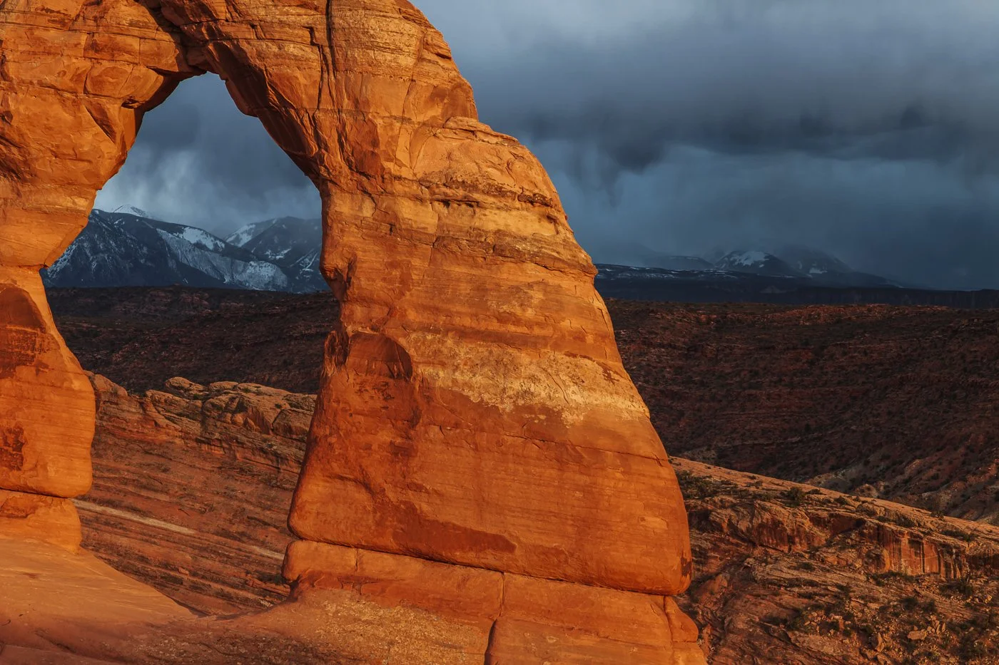 A close-up, tight crop of the right leg and opening of Delicate Arch in Arches National Park, glowing with warm orange light against a backdrop of dark, brooding storm clouds and snow-capped La Sal Mountains in the distance.