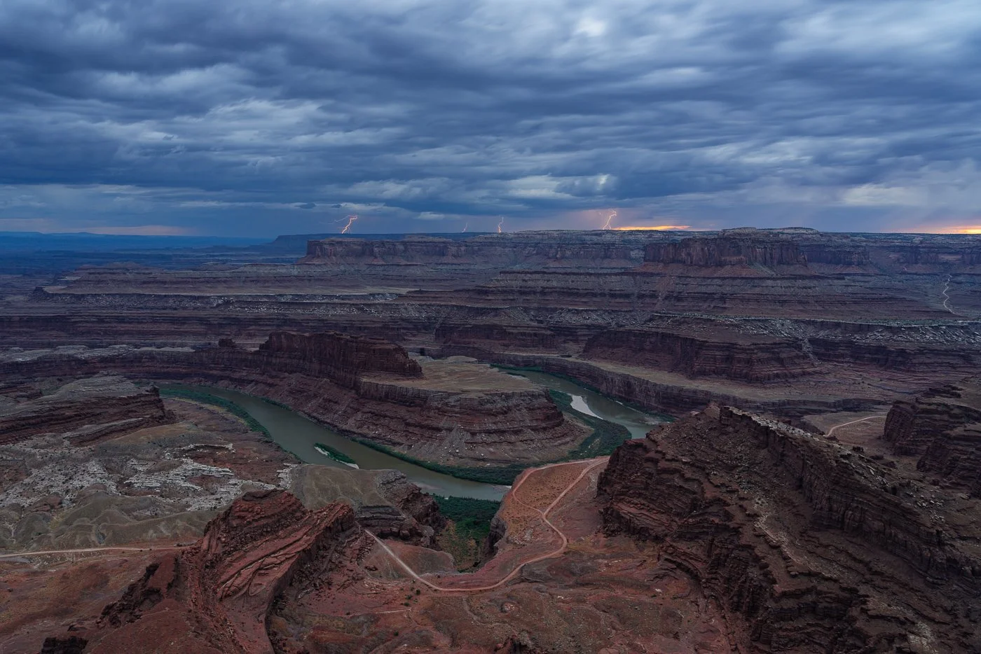 An expansive high-angle view overlooking the iconic gooseneck bend of the Colorado River at Dead Horse Point State Park under dark monsoon clouds, with multiple lightning bolts striking the distant canyon rim along the horizon.