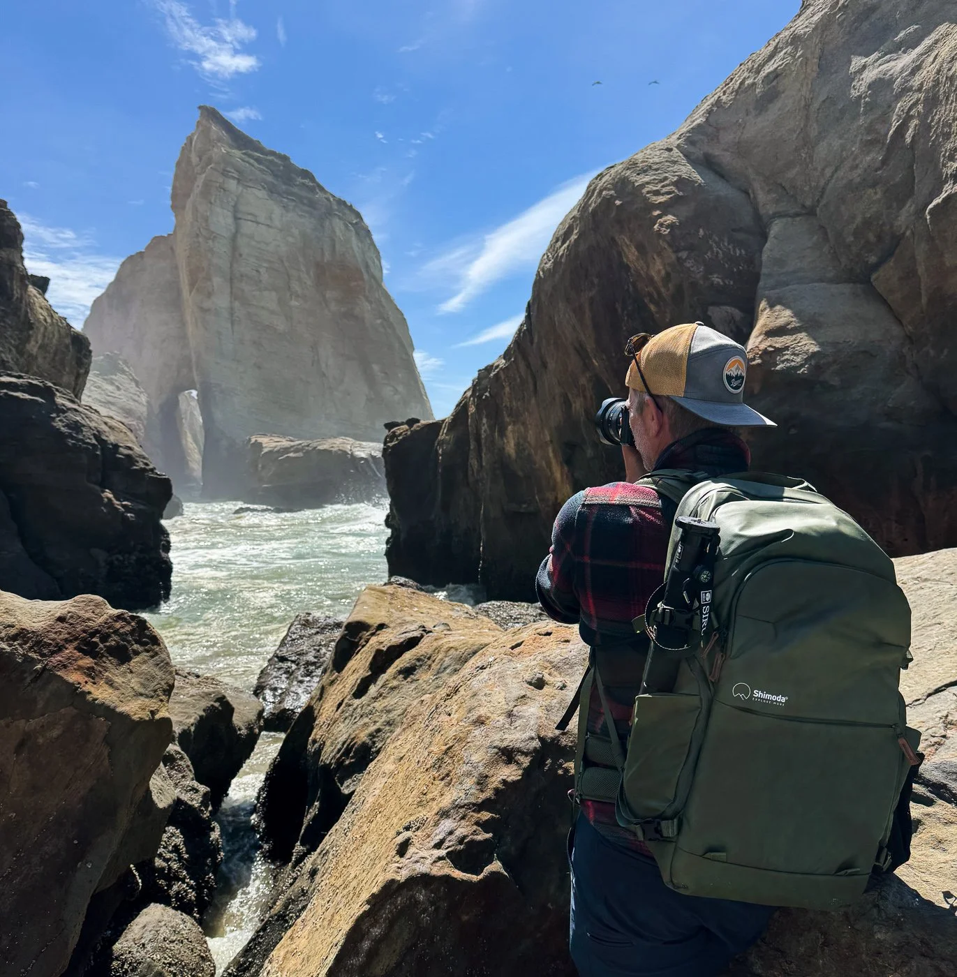 A photographer wearing a green Shimoda Explore v2 35 backpack stands on rugged coastal rocks, taking a photo of the sea stack at Cape Kiwanda on the Oregon Coast.