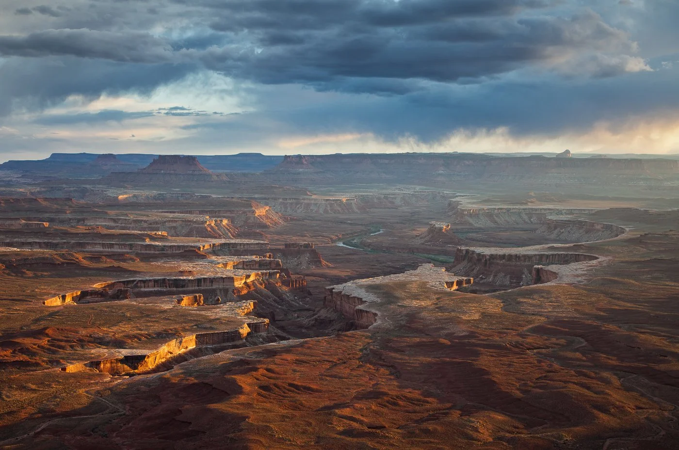 A high-angle, expansive landscape vista of the deeply carved canyons and white rim sandstone at Green River Overlook in Canyonlands National Park under a dramatic, stormy sky with sunset light illuminating the canyon edges.