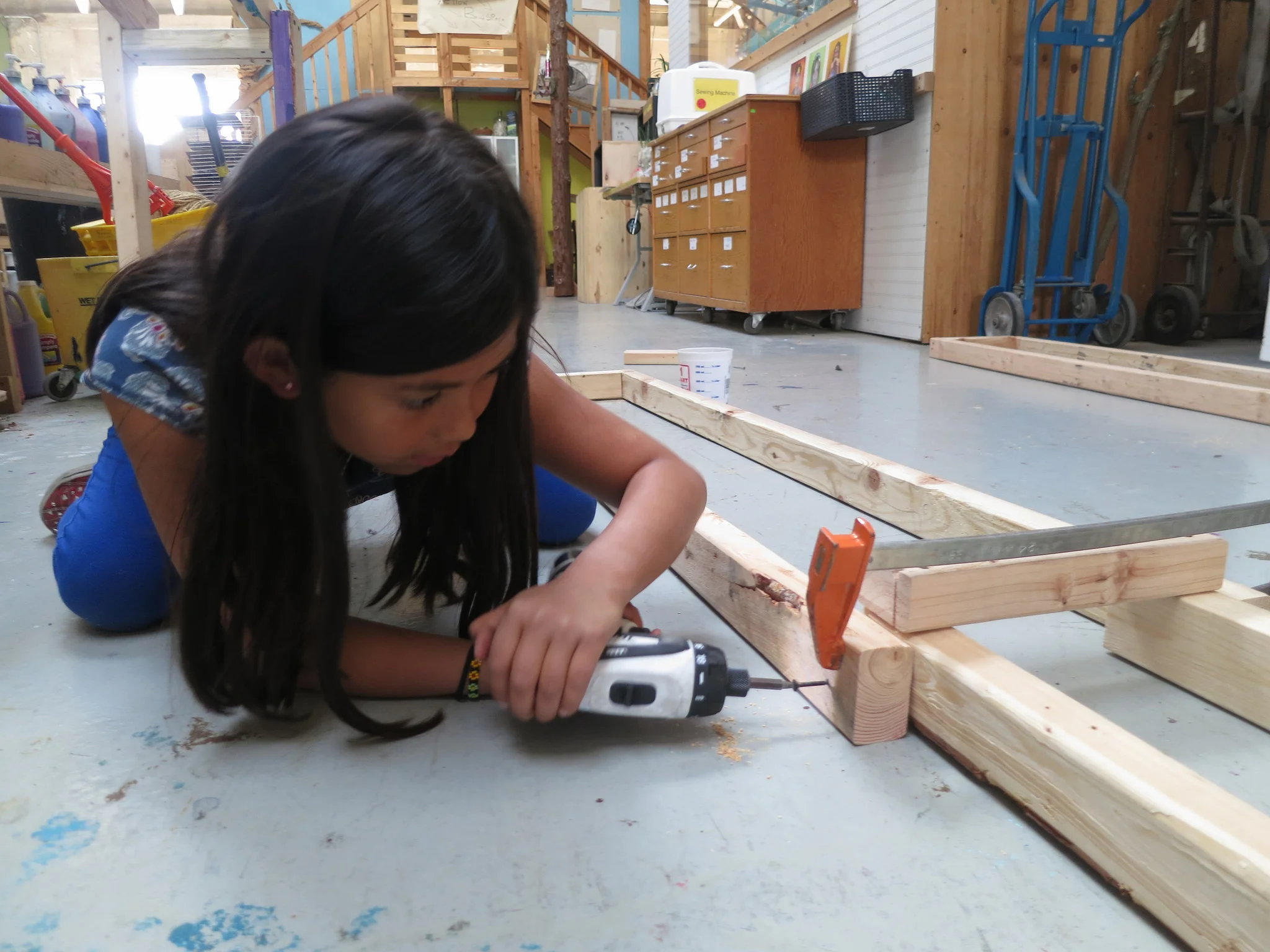 Maite grabs a drill and gets to work helping attach all the vertical ramp legs using the system established last week of driving only a single screw so they can be rotated upwards before being secured into place. 