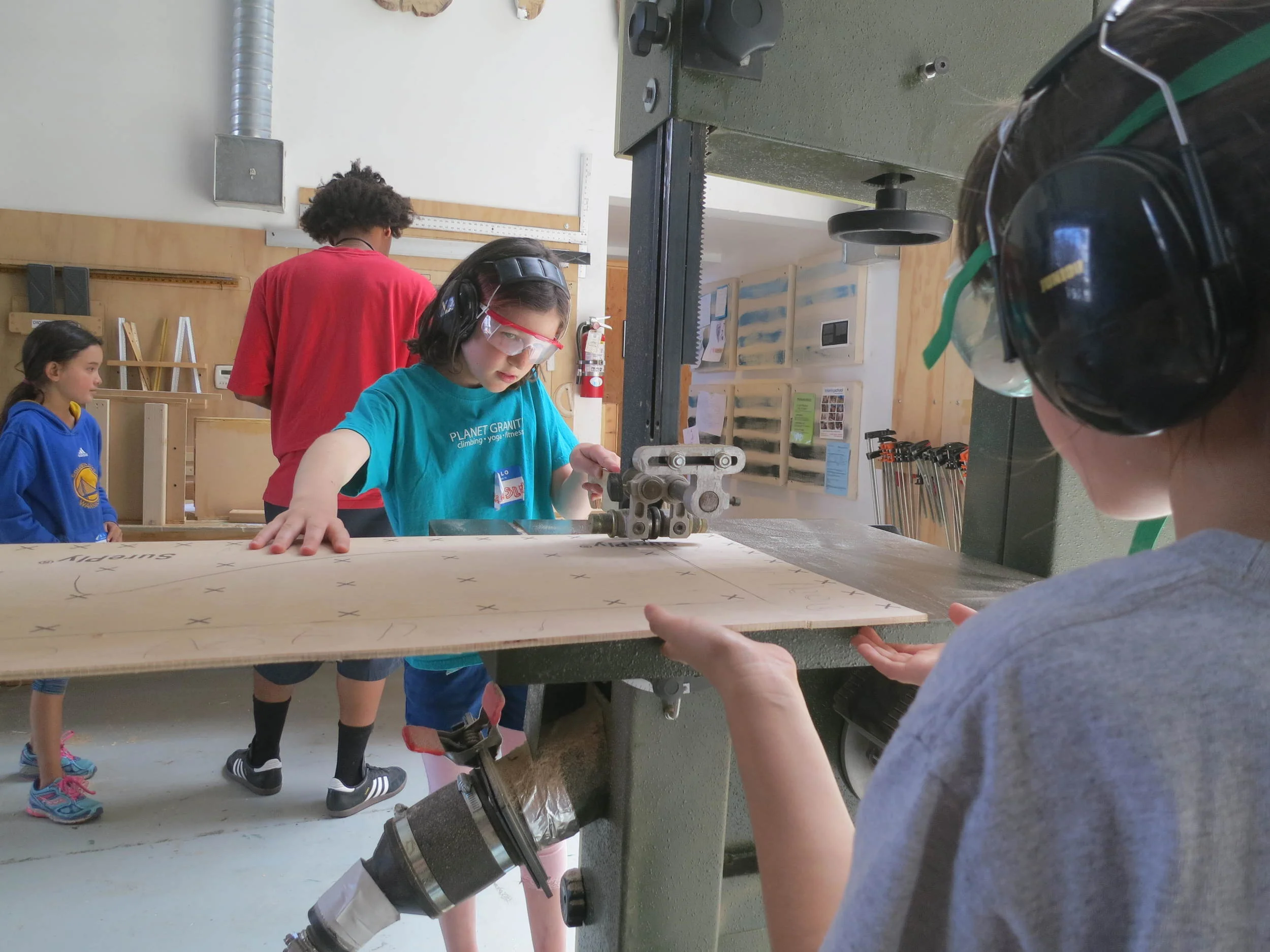 Ainsley focuses on lining up the bandsaw cut. The wood will later help sheath the toll booth. Julia assists by making sure the cut wood stays parallel to the floor. 