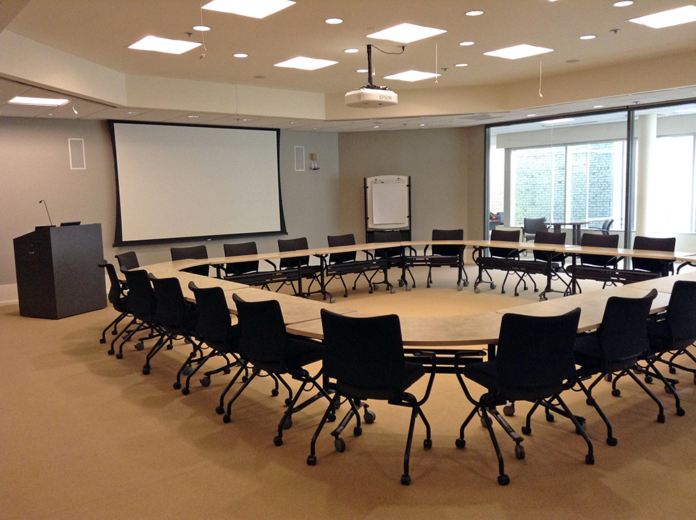 Large room with several chairs around a circle of tables, a screen and podium in the front of the room.