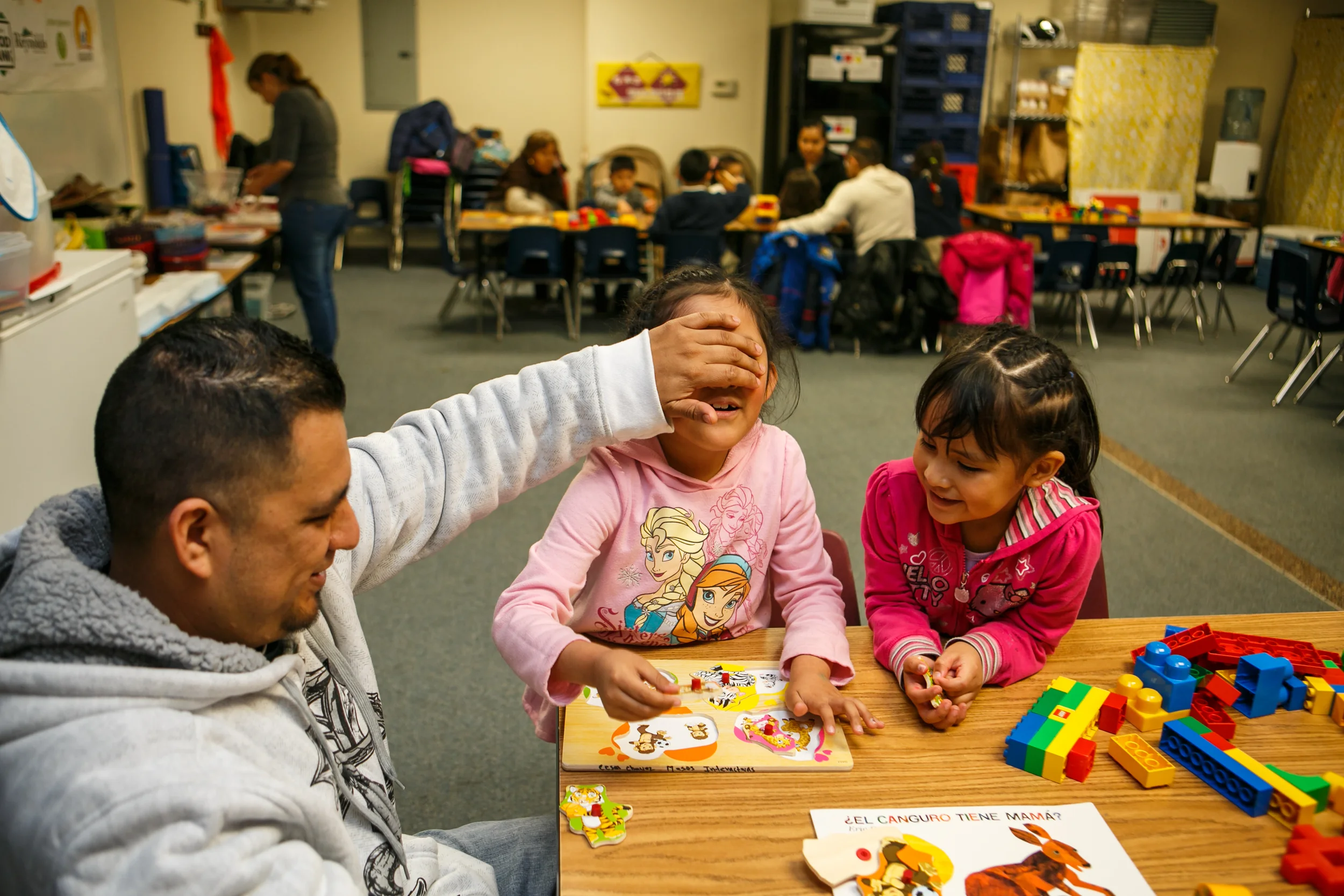 Father sitting in a classroom with two children, playing with a puzzle.