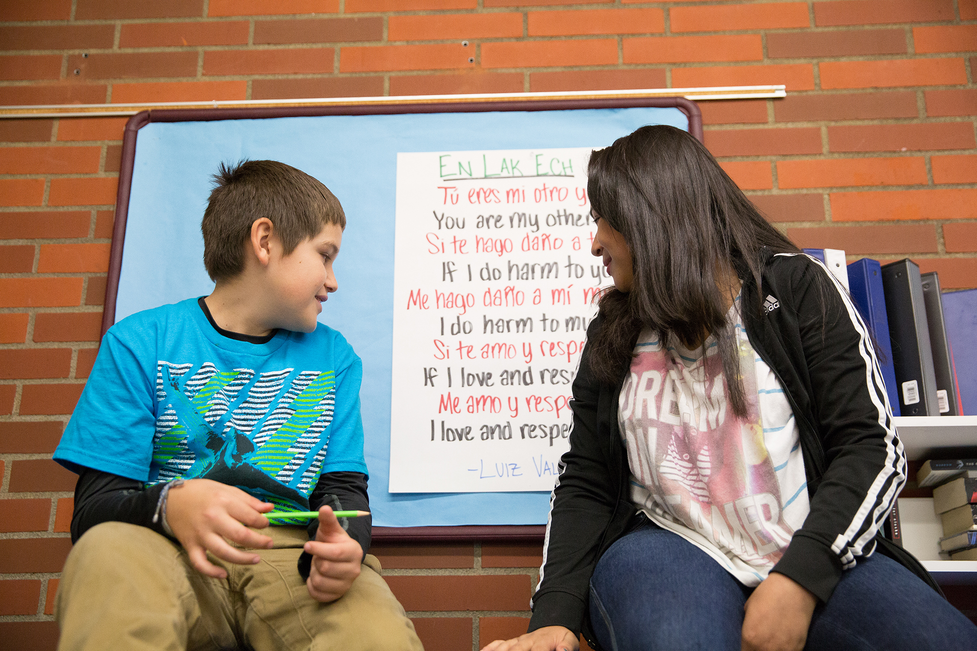 Two youth sitting on a table, looking over their shoulders at a poster with a poem in two languages.