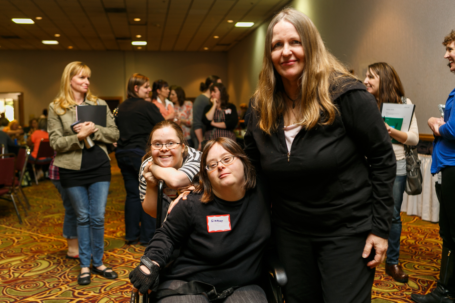 Three women pose for the camera in a crowded conference room. Two appear to have Down Syndrome; one is in a wheelchair.