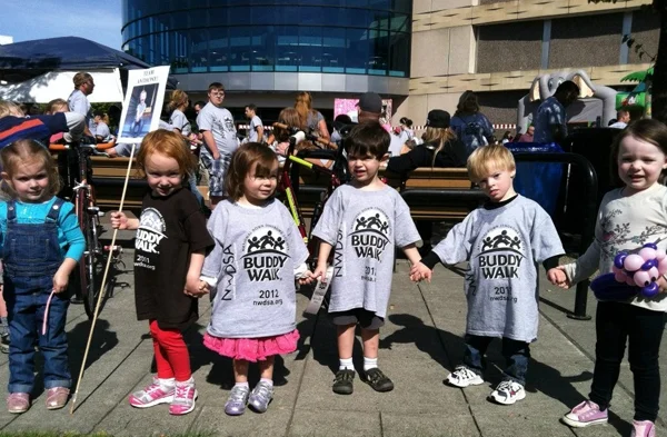 A row of children in oversized t-shirts printed with the words "Buddy Walk" hold hands.