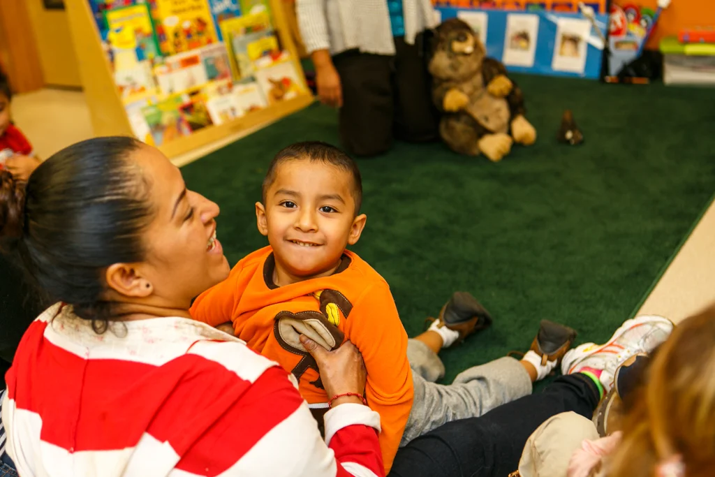A woman sits on the floor holding her child. He looks over her shoulder, smiling. She is laughing. 