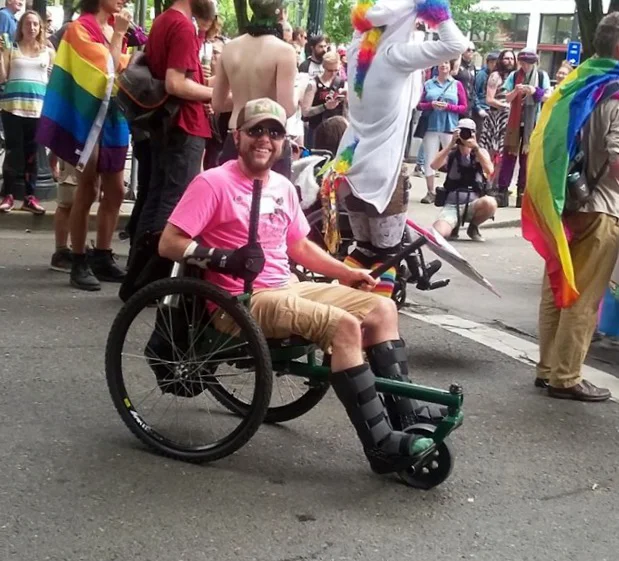 Nico sits in their GRIT Freedom Chair in the middle of a street and smiles. Behind them people wear rainbow flag capes and a unicorn hoodie.