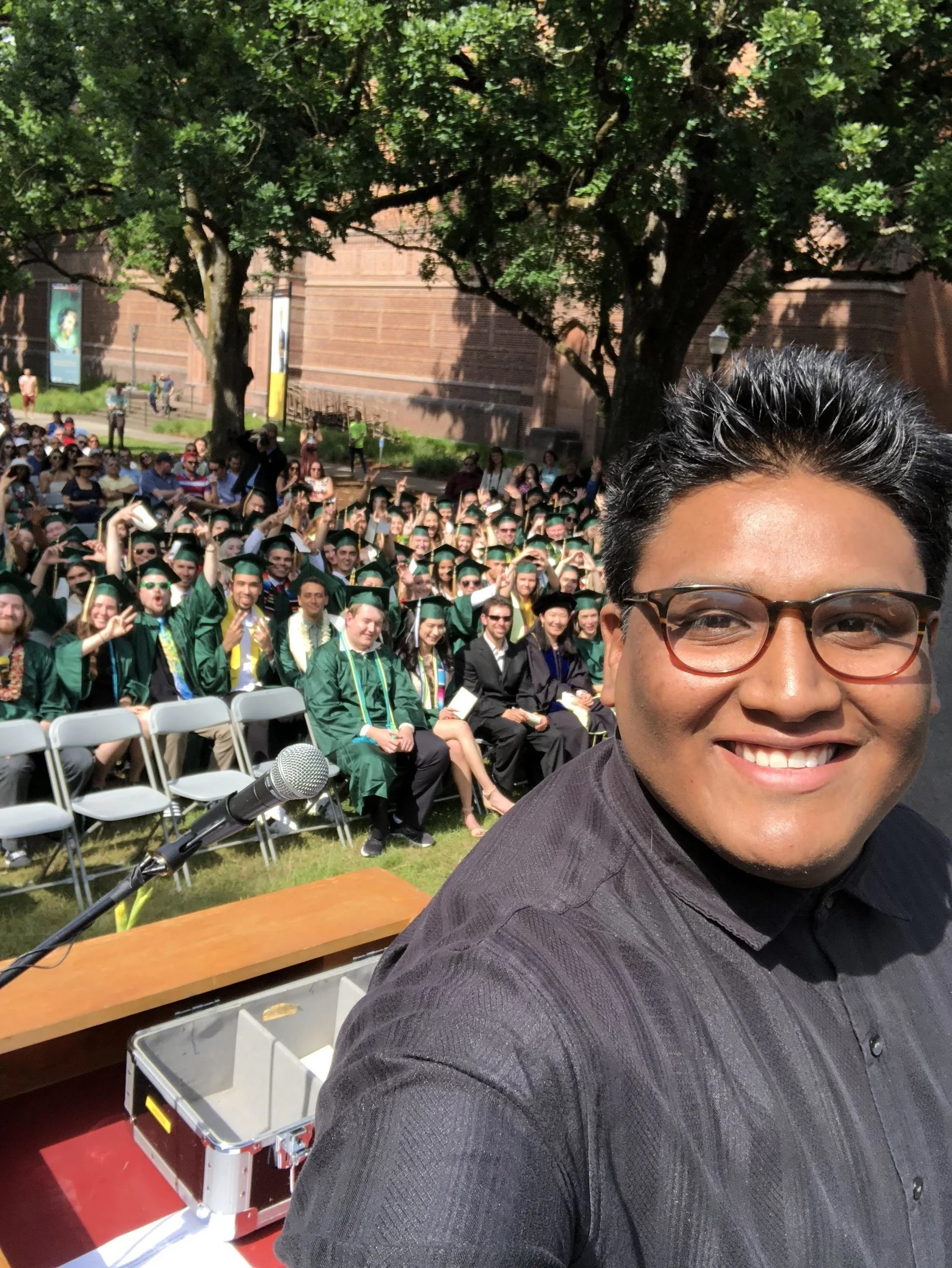 Joel stands on stage smiling with a group of graduates in caps and gowns seated behind him, facing the stage.