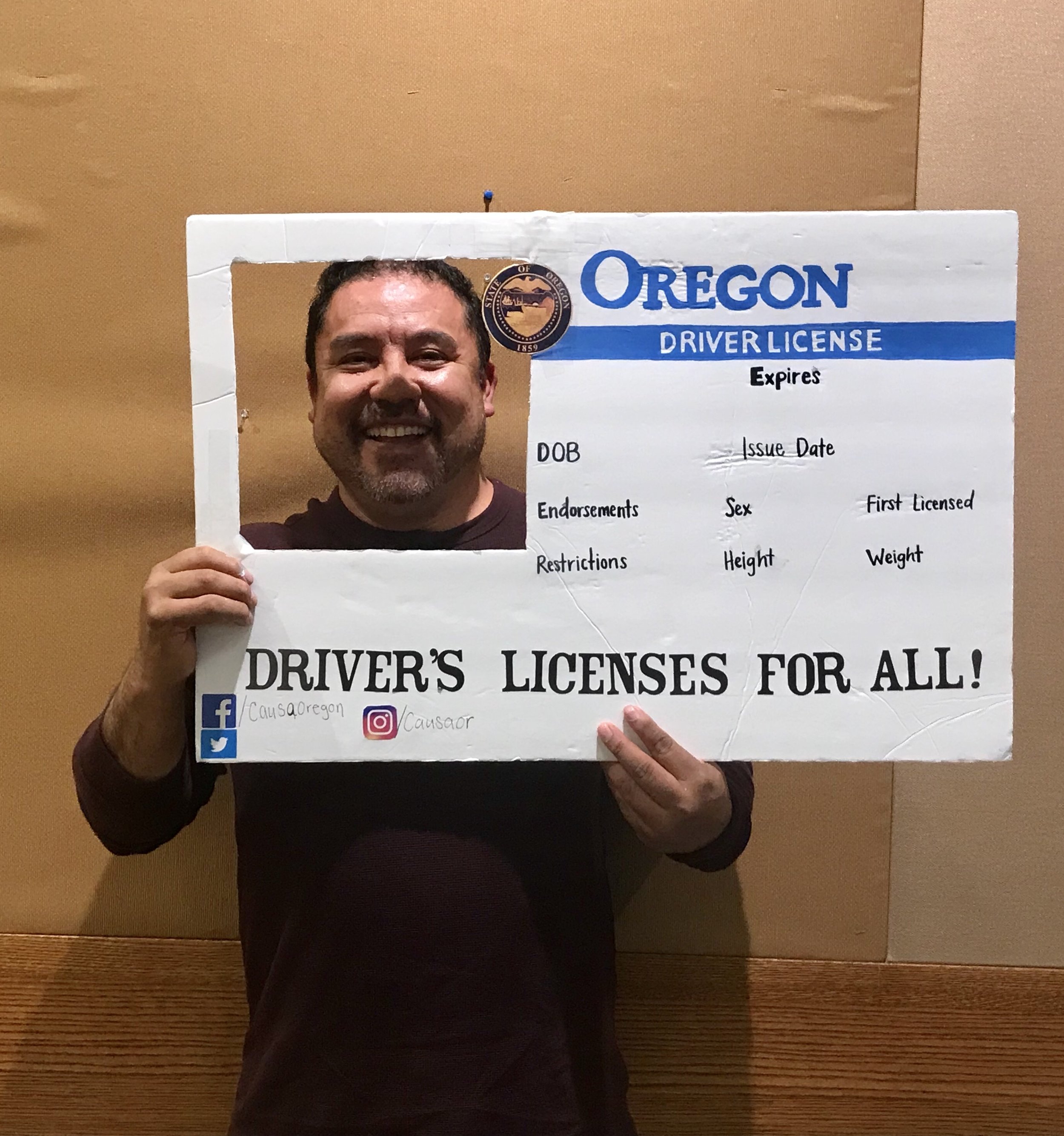 A parent holds up a poster decorated to look like a giant Oregon driver’s license. His face is framed by a square cut-out in the upper left corner.