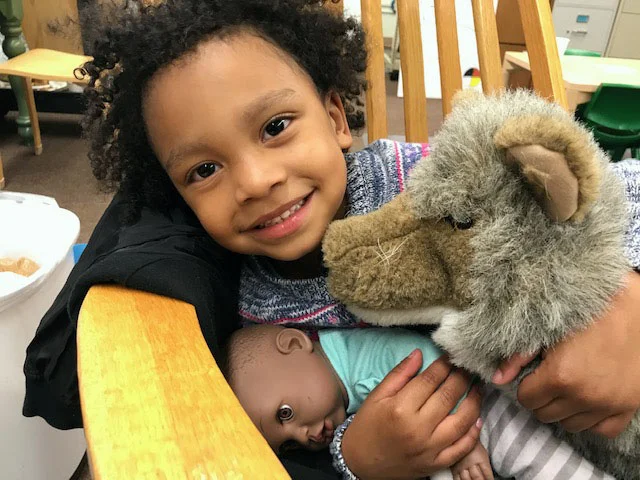 A smiling child curls up with a babydoll and a stuffed animal.