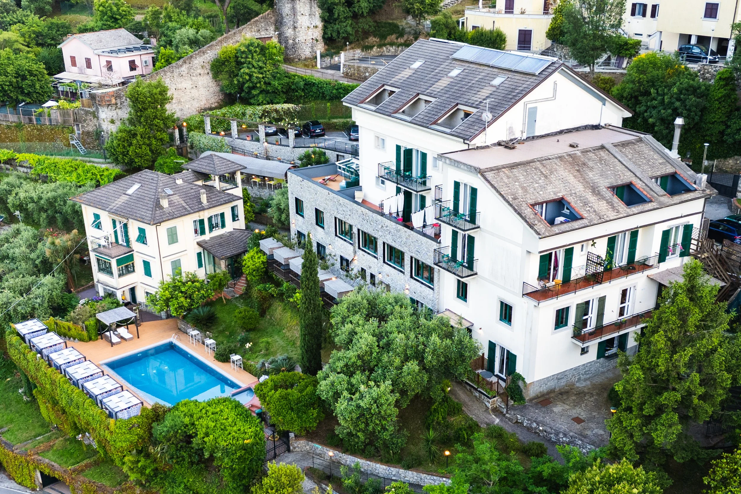 Aerial view of a large multi-story white apartment building with balconies, green shutters, and rooftop skylights. The building is surrounded by lush green trees and gardens, including a swimming pool with chairs and tables nearby. A smaller building and other houses are visible in the surrounding neighborhood.