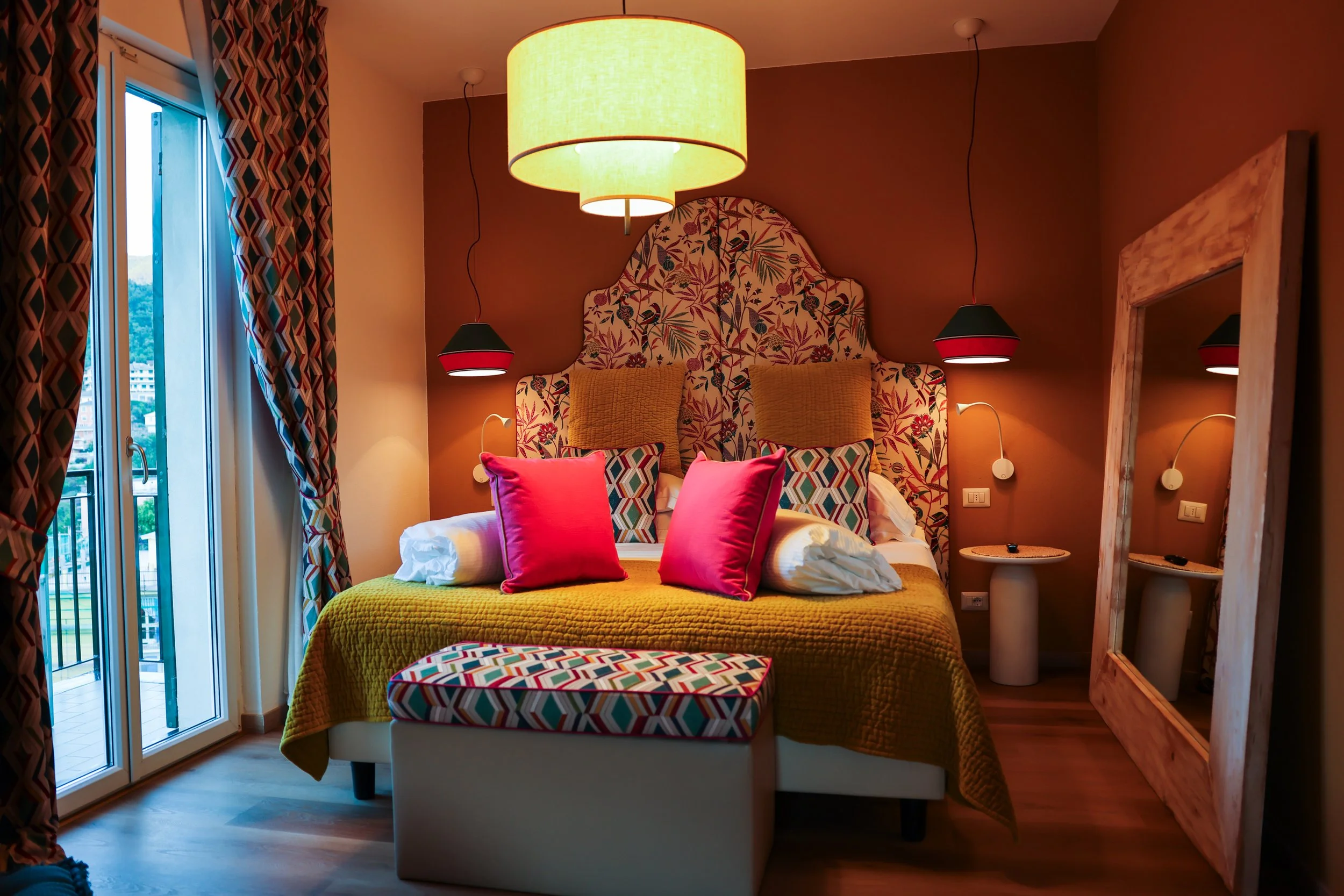 A cozy hotel bedroom with a large bed featuring a colorful tufted headboard, bright pink and patterned pillows, and a yellow bedspread. There is a large round ceiling lamp, two black-and-red pendant lights, and two wall-mounted reading lamps. To the right, a full-length mirror leans against the wall, and near the bed, a door with patterned curtains leads to a balcony.