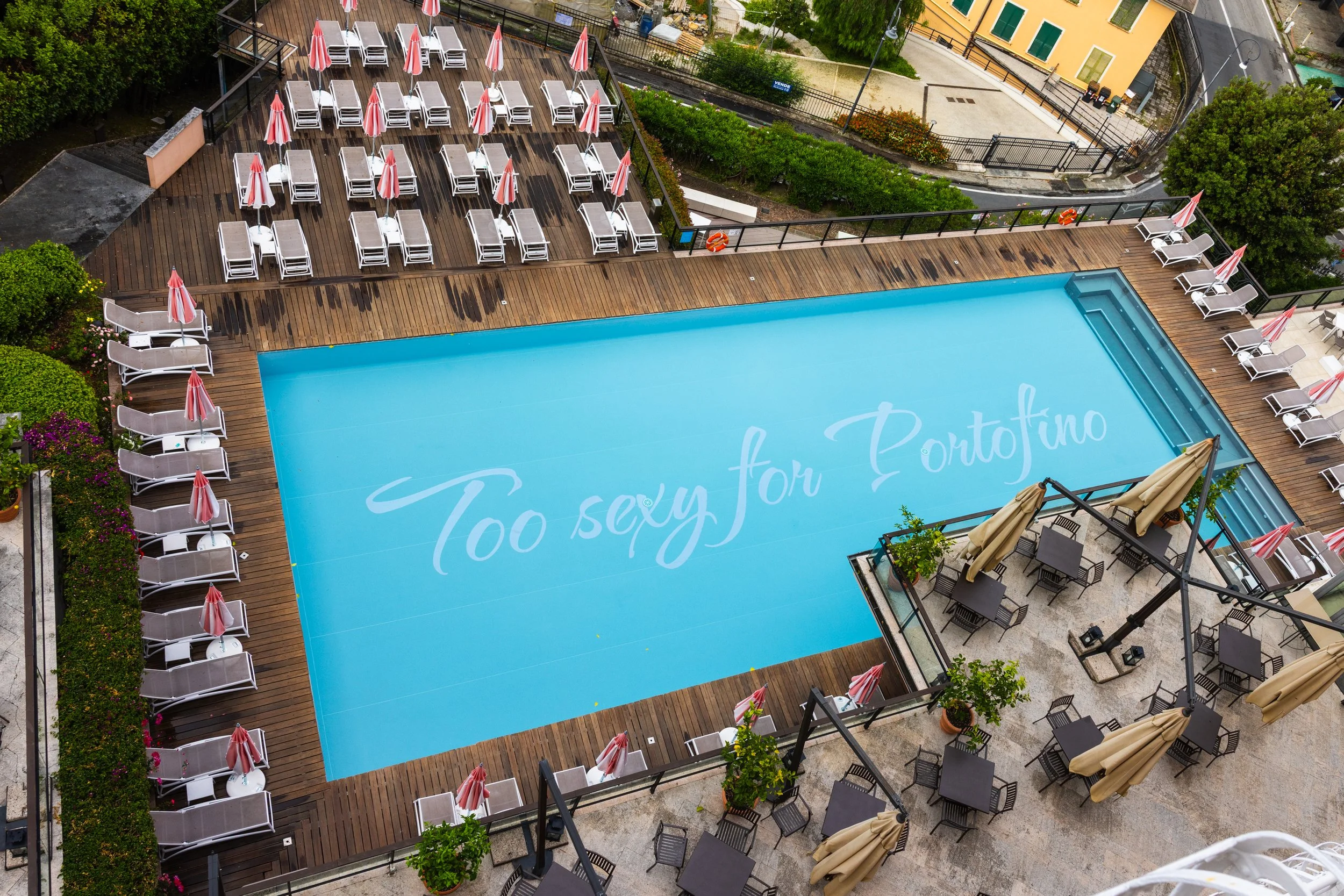 An aerial view of a swimming pool with the words 'Too sexy for Portofino' written on the pool surface. The pool is surrounded by a wooden deck with lounge chairs, umbrellas, and some tables with umbrellas. There are also some potted plants around the area.
