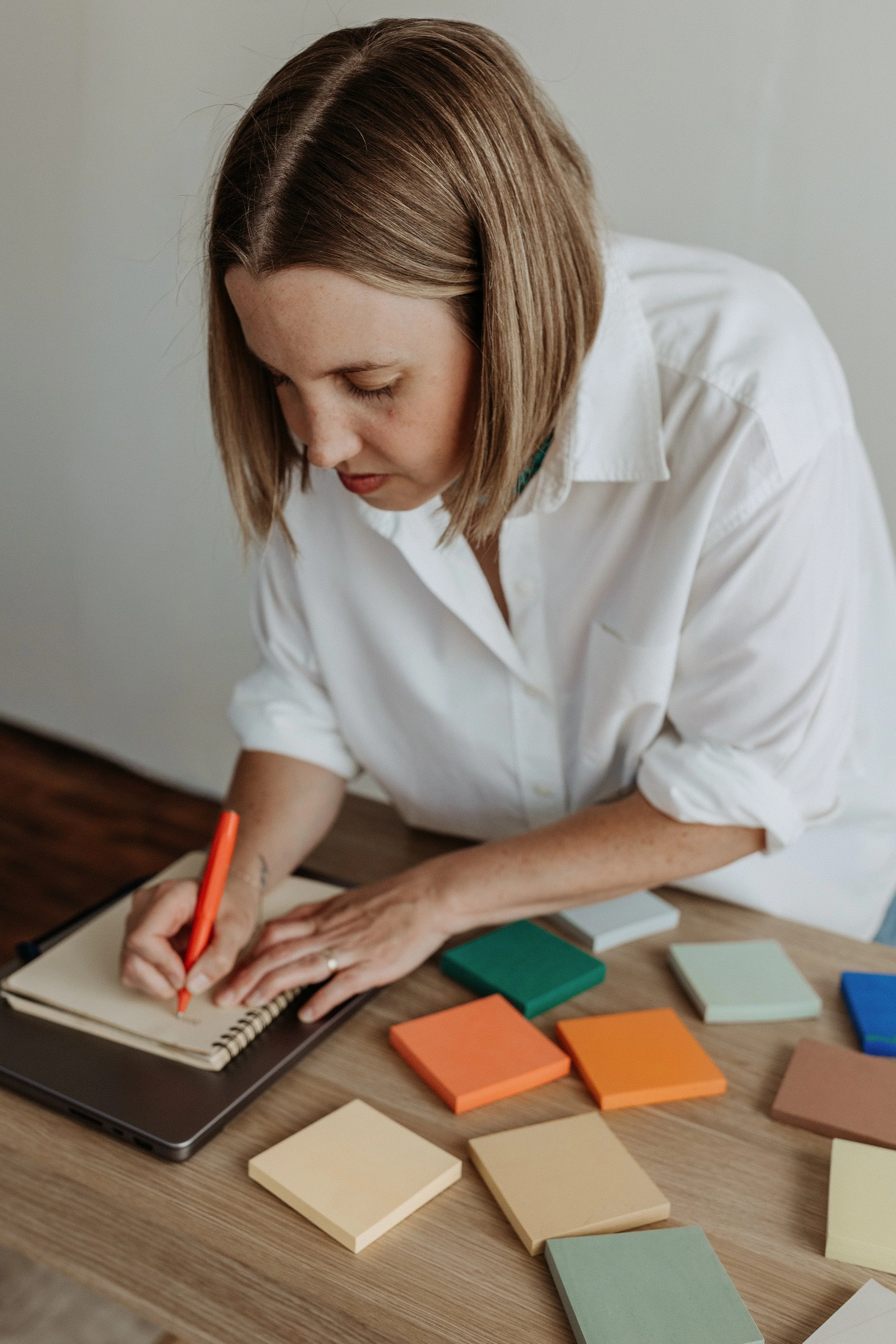 Woman with brown hair writing in a notebook on a wooden desk surrounded by color swatches.