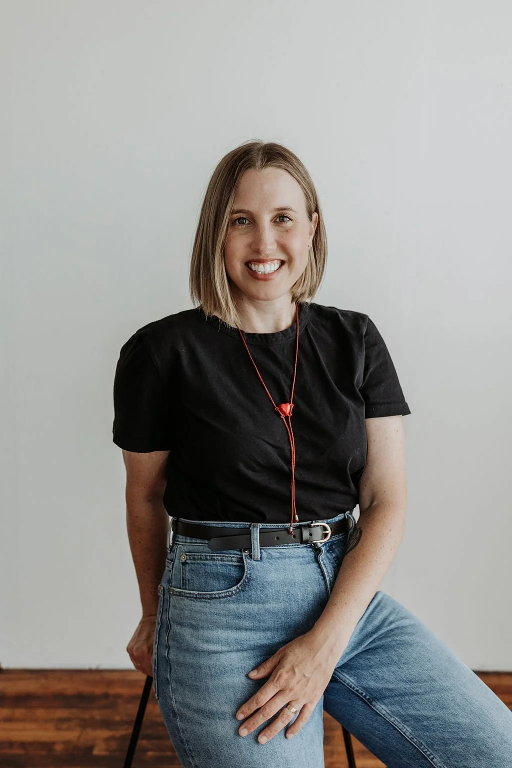 A woman with shoulder-length blonde hair smiling, wearing a black t-shirt, light blue jeans, and a red heart-shaped locket necklace, seated against a plain white wall.
