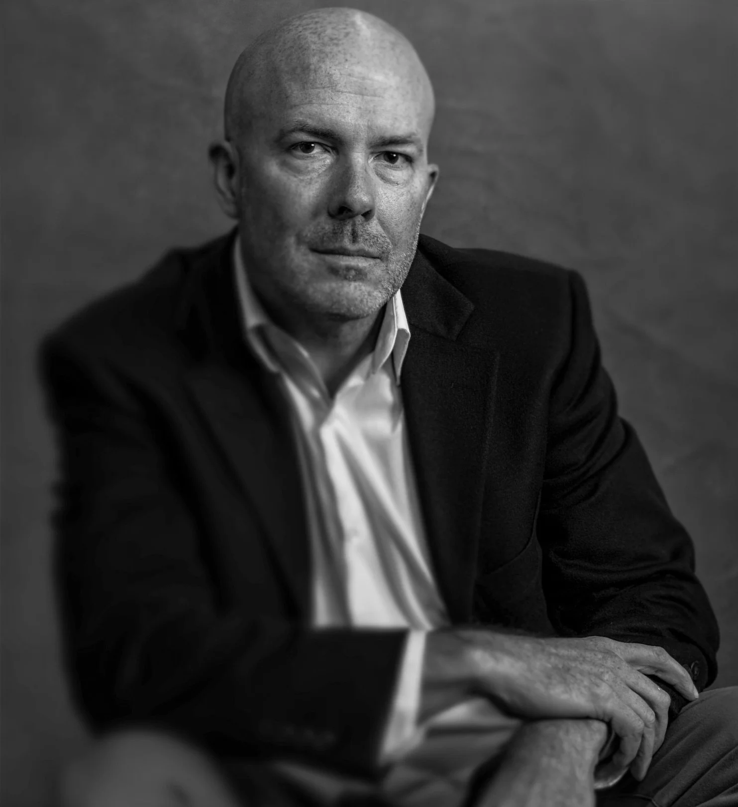 A black and white portrait of a middle-aged man with a bald head, wearing a suit and white shirt, sitting with one arm resting on his lap, looking directly at the camera with a serious expression.