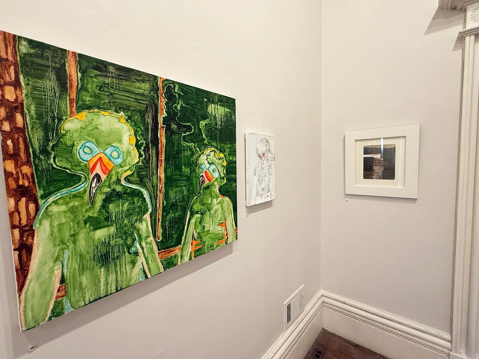 Gallery wall with four art pieces: a large green abstract painting of two figures wearing blue goggles and orange masks, a smaller black and white portrait of a child, and a framed photograph of a person wearing a striped shirt standing in front of a