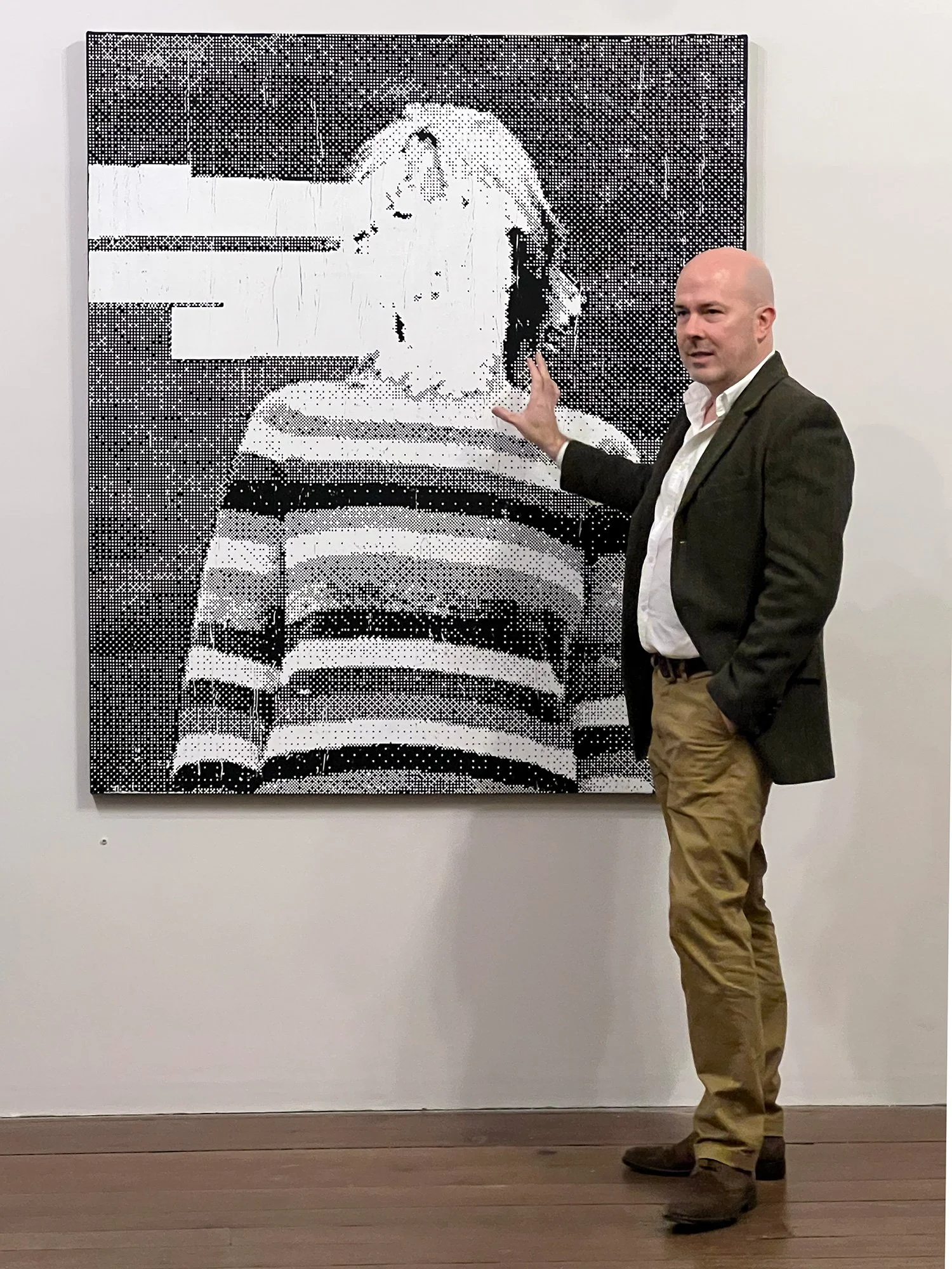 A man in a white shirt and dark blazer standing next to a black-and-white pixelated portrait of a woman with short hair, wearing a striped top, in an art gallery.