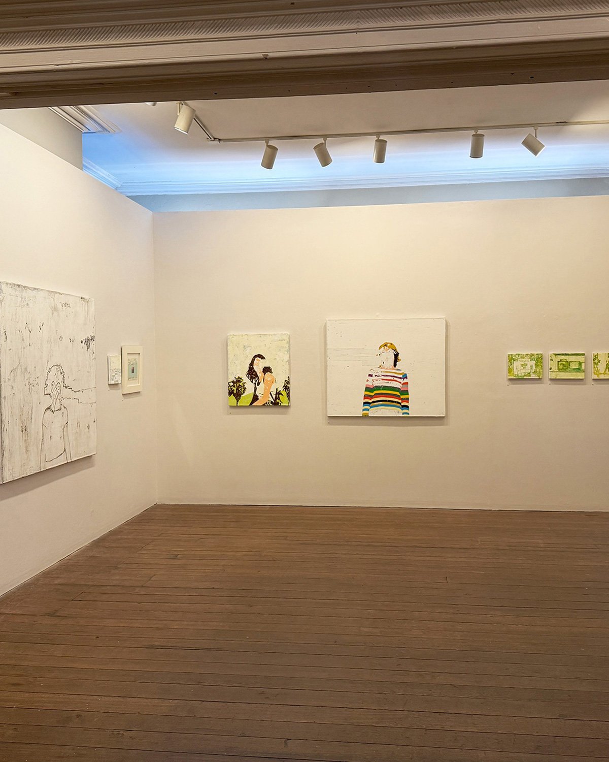 Art gallery with white walls, wooden floor, and several paintings including sketches and colorful portraits, with ceiling track lighting.
