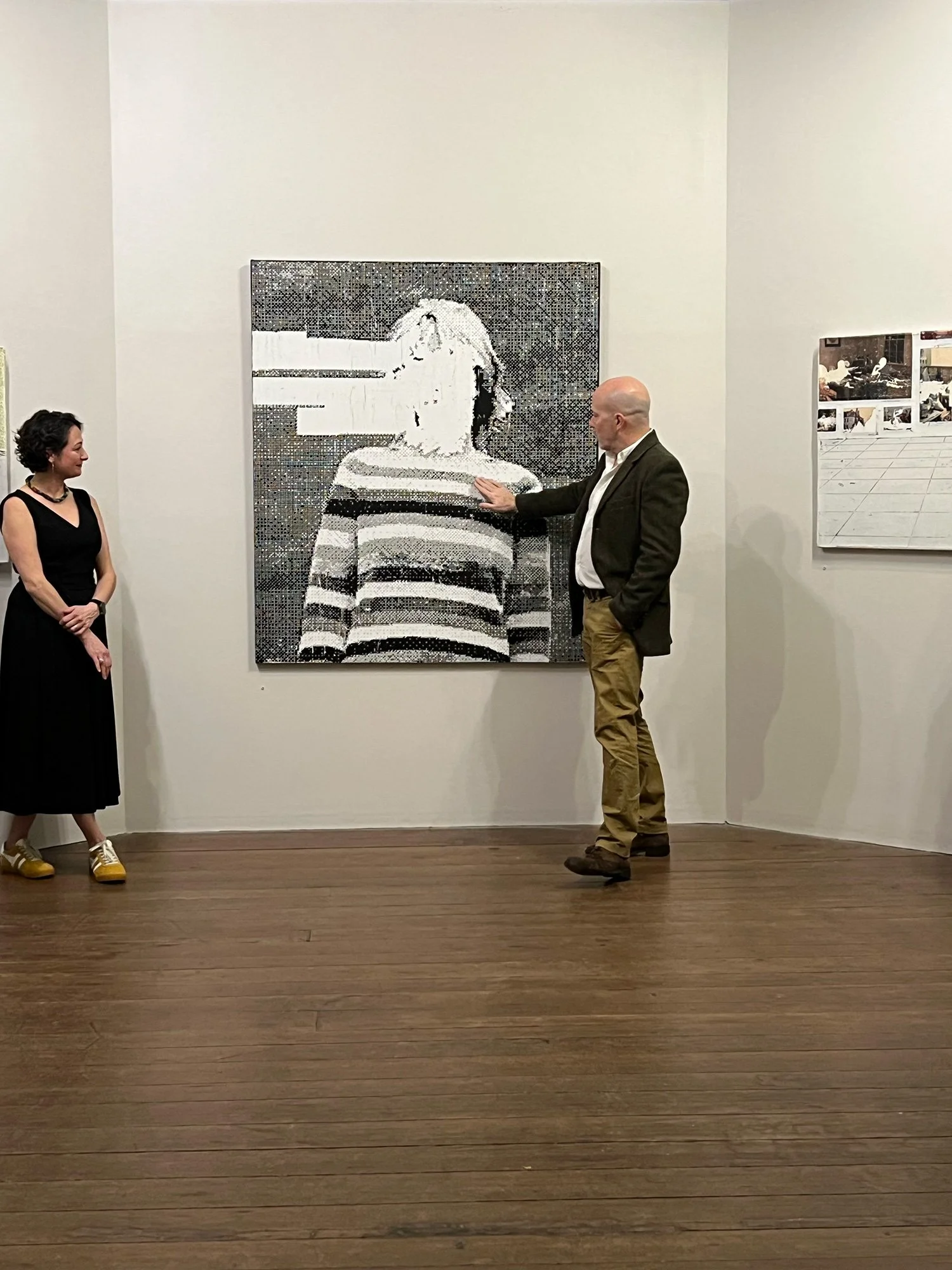A man and a woman are viewing a mosaic portrait of a woman in an art gallery. The portrait is composed of small tiles, and the man is pointing at it.