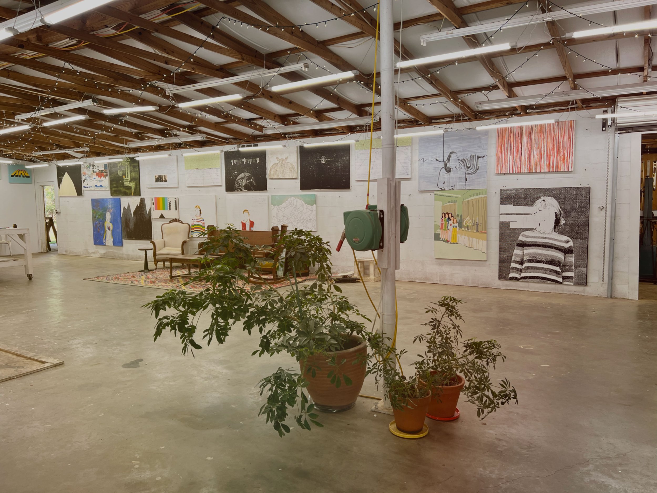 Art gallery with paintings on the wall, plants in pots, string lights on the ceiling, and vintage chairs and a sofa on the floor.