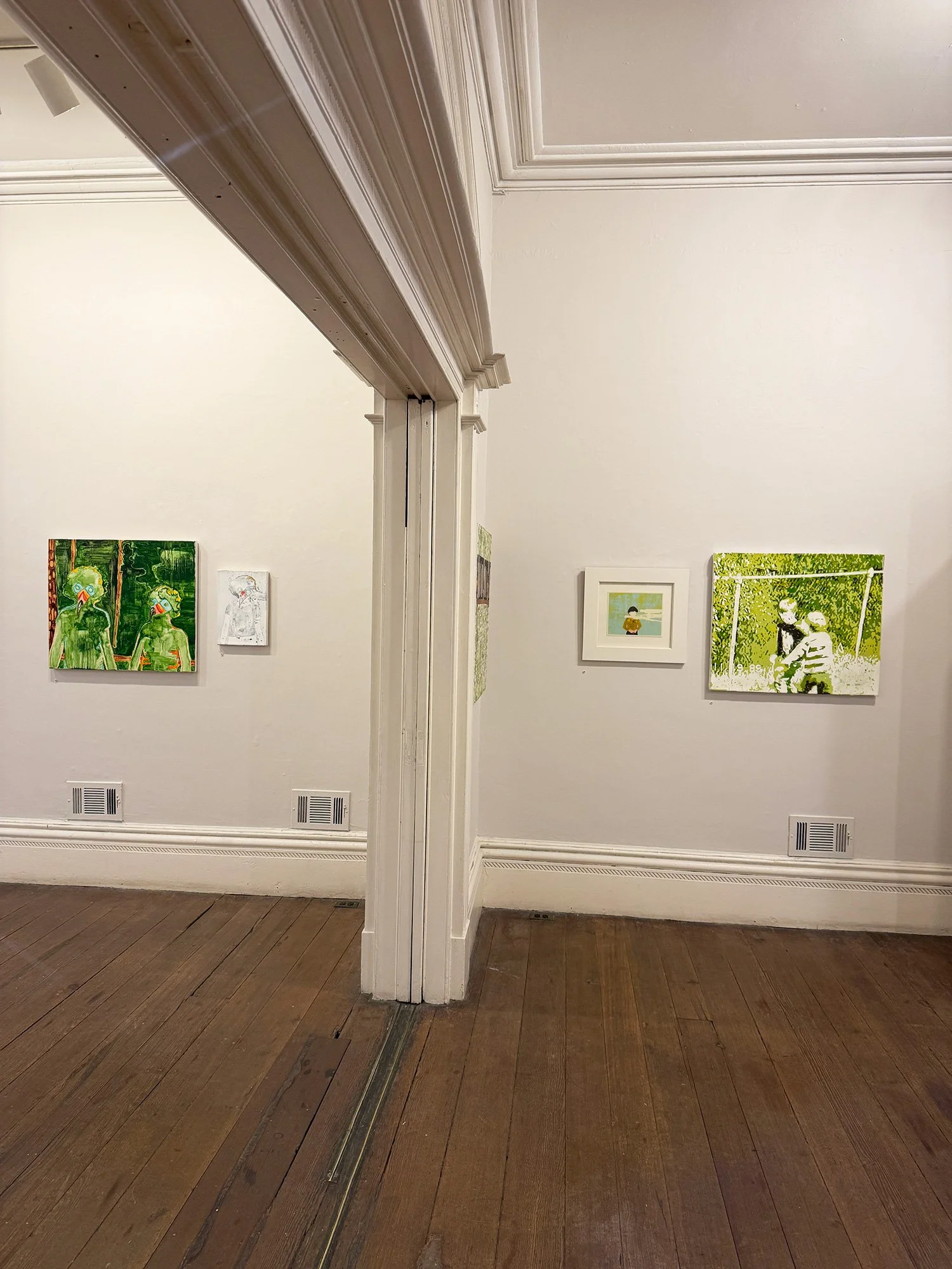 An art gallery with white walls and wood flooring, featuring several colorful paintings. The image is taken from the corner where two walls meet.
