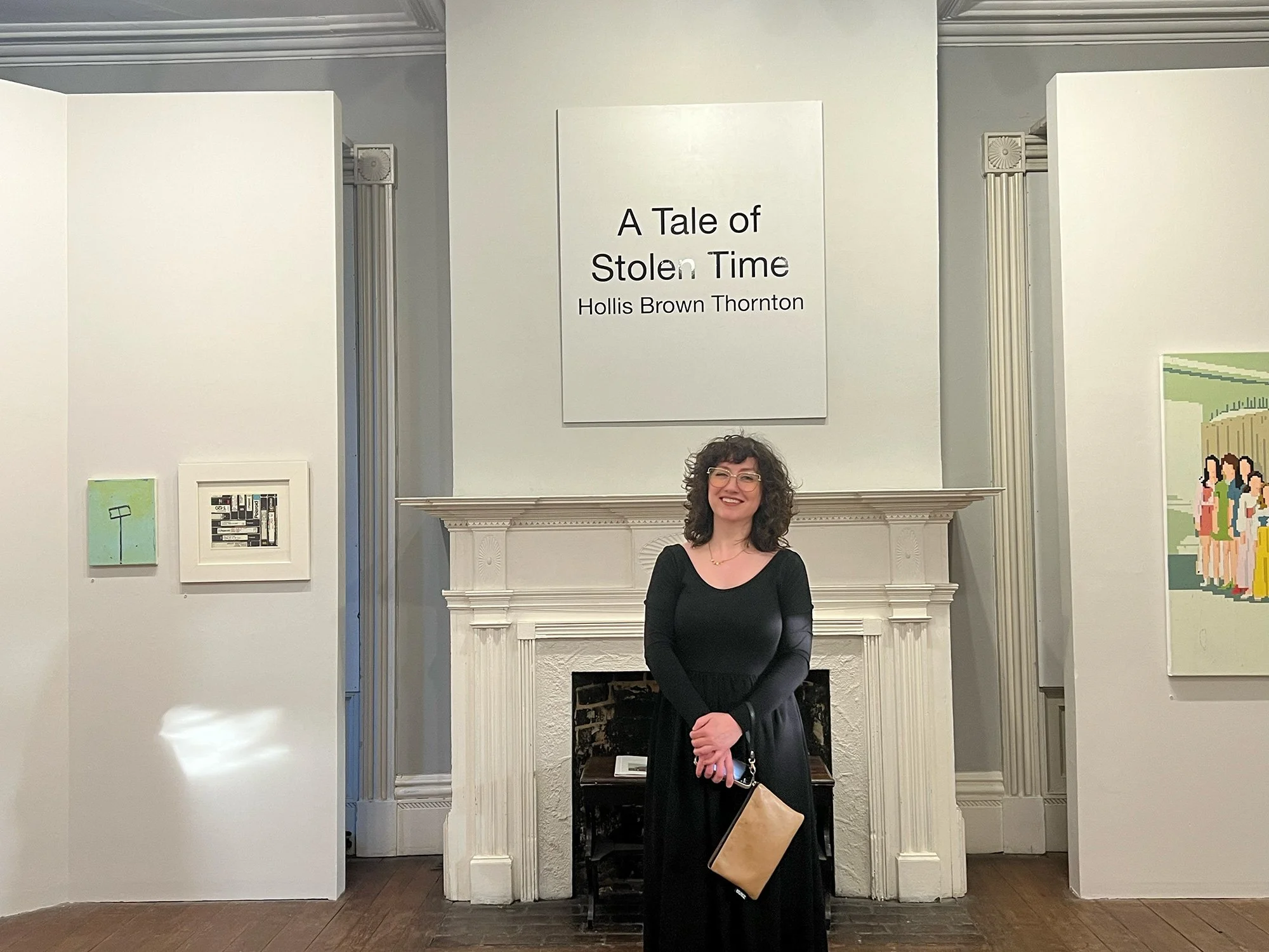 Woman with curly hair and glasses standing in front of a white fireplace, smiling, in an art gallery. There is a sign above the fireplace that reads, 'A Tale of Stolen Time, Hollis Brown Thornton.' Artworks are displayed on the walls to her sides.