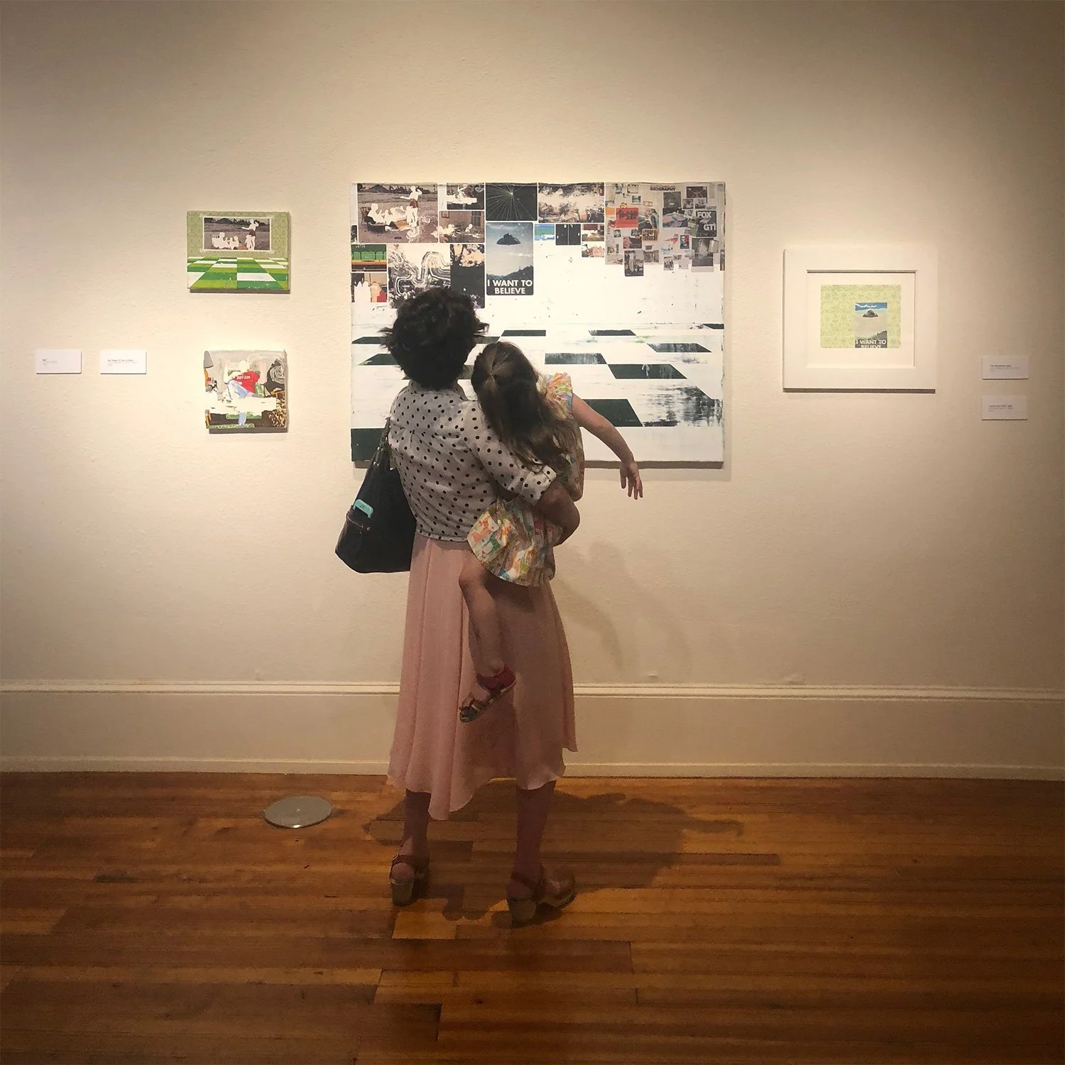 Visitor holding a child while viewing an exhibition of work by Hollis Brown Thornton, with paintings, drawings, and mixed imagery displayed on the gallery wall.