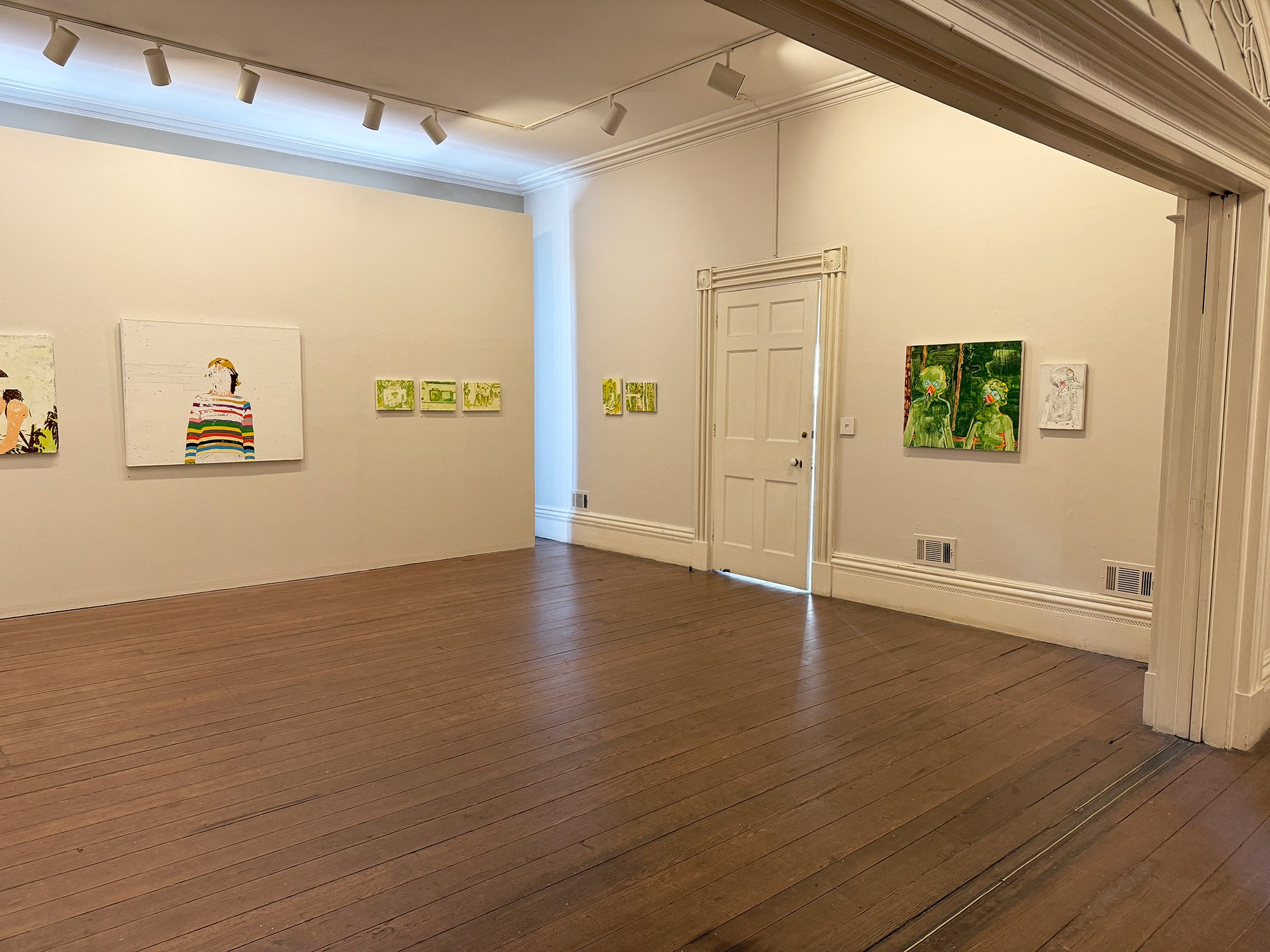 An art gallery with white walls displaying various paintings, a wooden floor, and a door at the back. Track lighting is mounted on the ceiling.