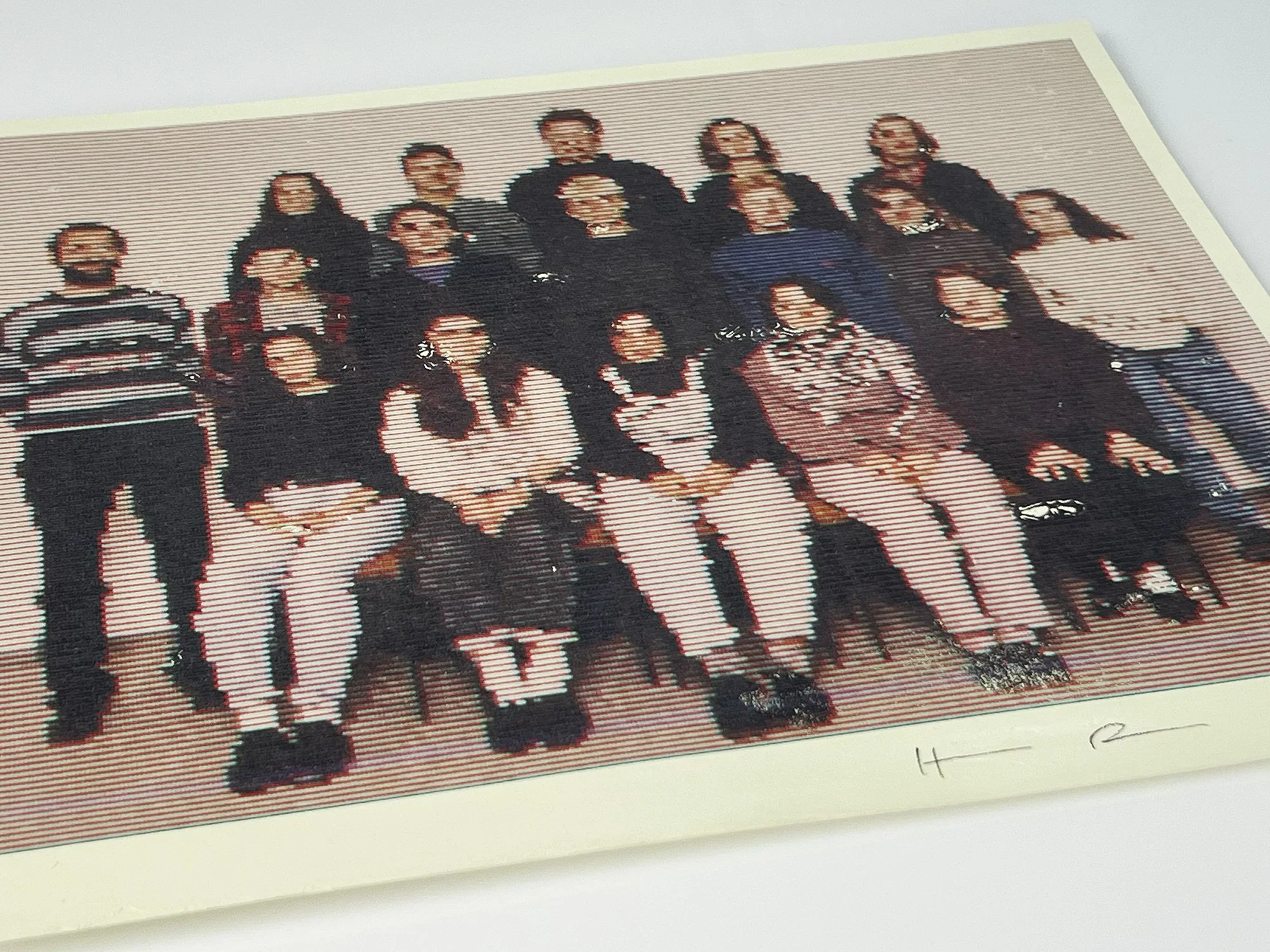 A cross-stitch artwork of eleven people standing together, with two kneeling in the front, some holding objects, all dressed casually.