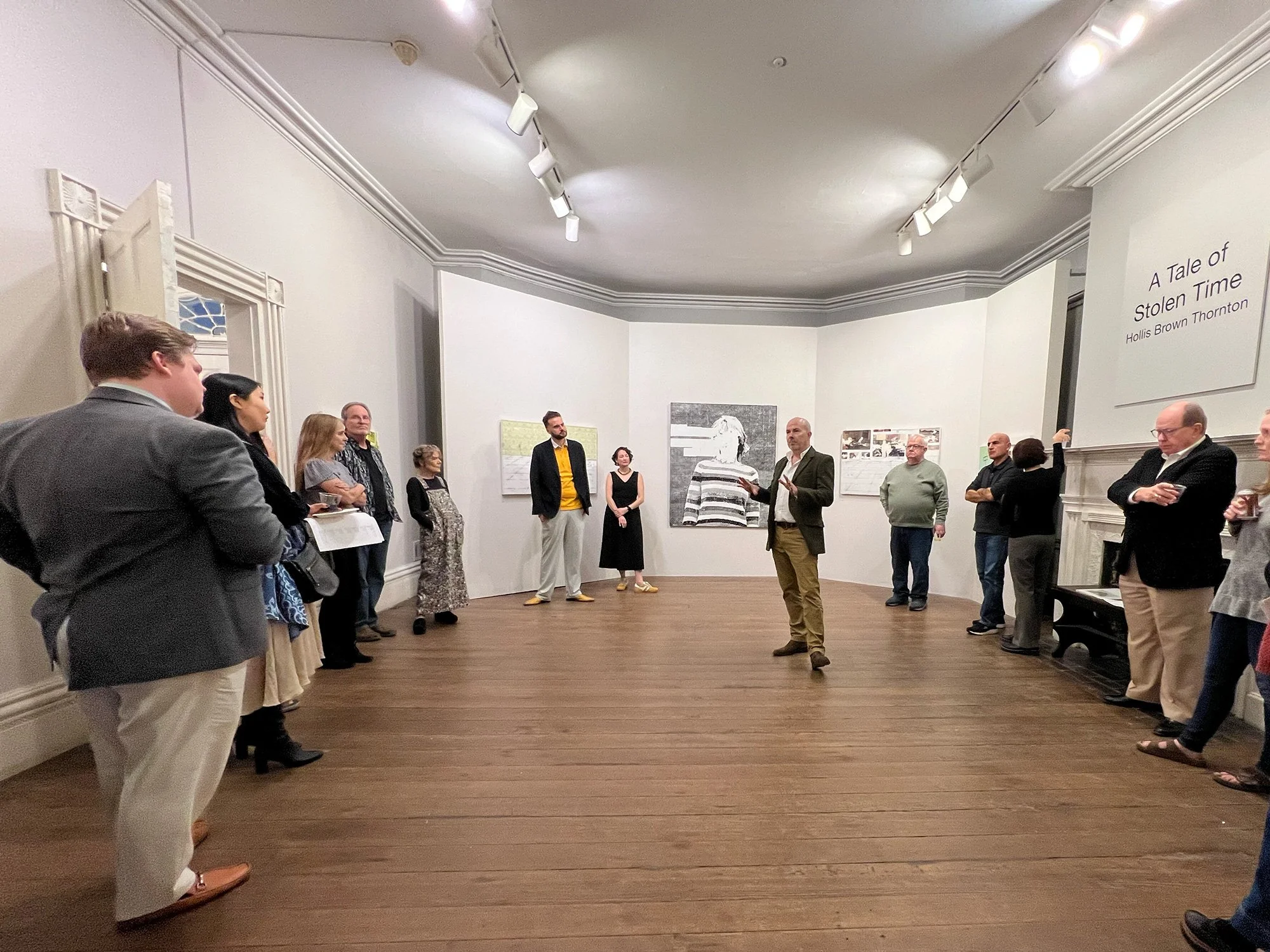 A group of people attending an art gallery tour, listening to a man in a blazer and khakis explain artwork. Art pieces are displayed on white walls in a spacious room with wooden floors.