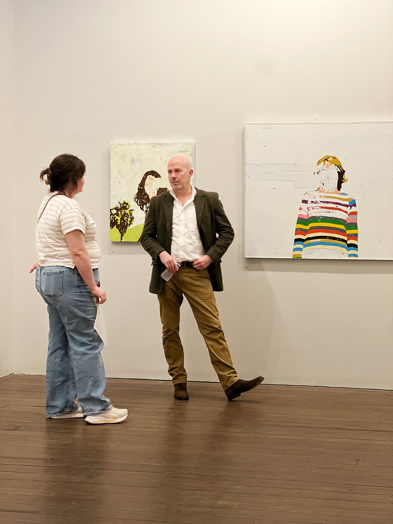 Two people, a woman in a striped shirt and jeans and a man in a blazer and khakis, are talking in an art gallery. There are three paintings on the wall behind them, including an abstract portrait of a woman and a colorful striped figure.