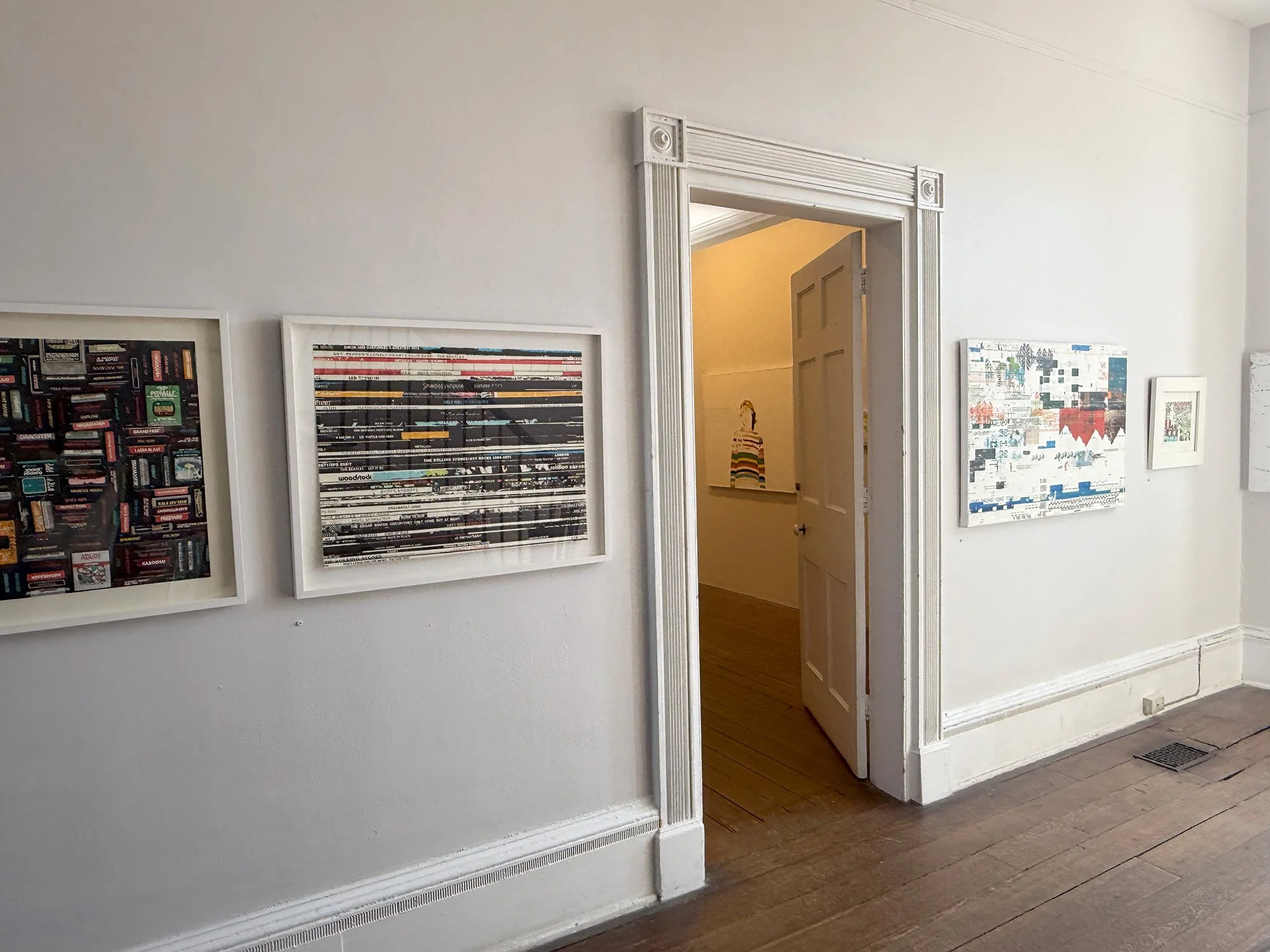 Art gallery with framed abstract and collage artworks on white walls, wooden floor, open doorway to another room with art.