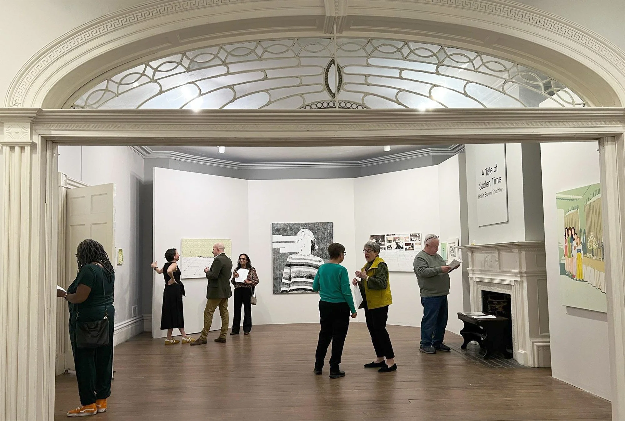 People viewing and discussing artwork in an art gallery with white walls, wooden floors, and decorative ceiling architecture.