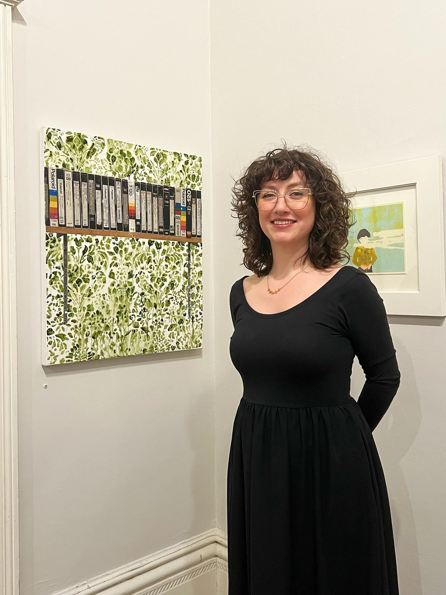 A woman with curly hair, glasses, wearing a black dress, standing in an art gallery, smiling at the camera. Behind her are two pieces of art, one with a leafy green pattern and a framed illustration of a person sitting outdoors.