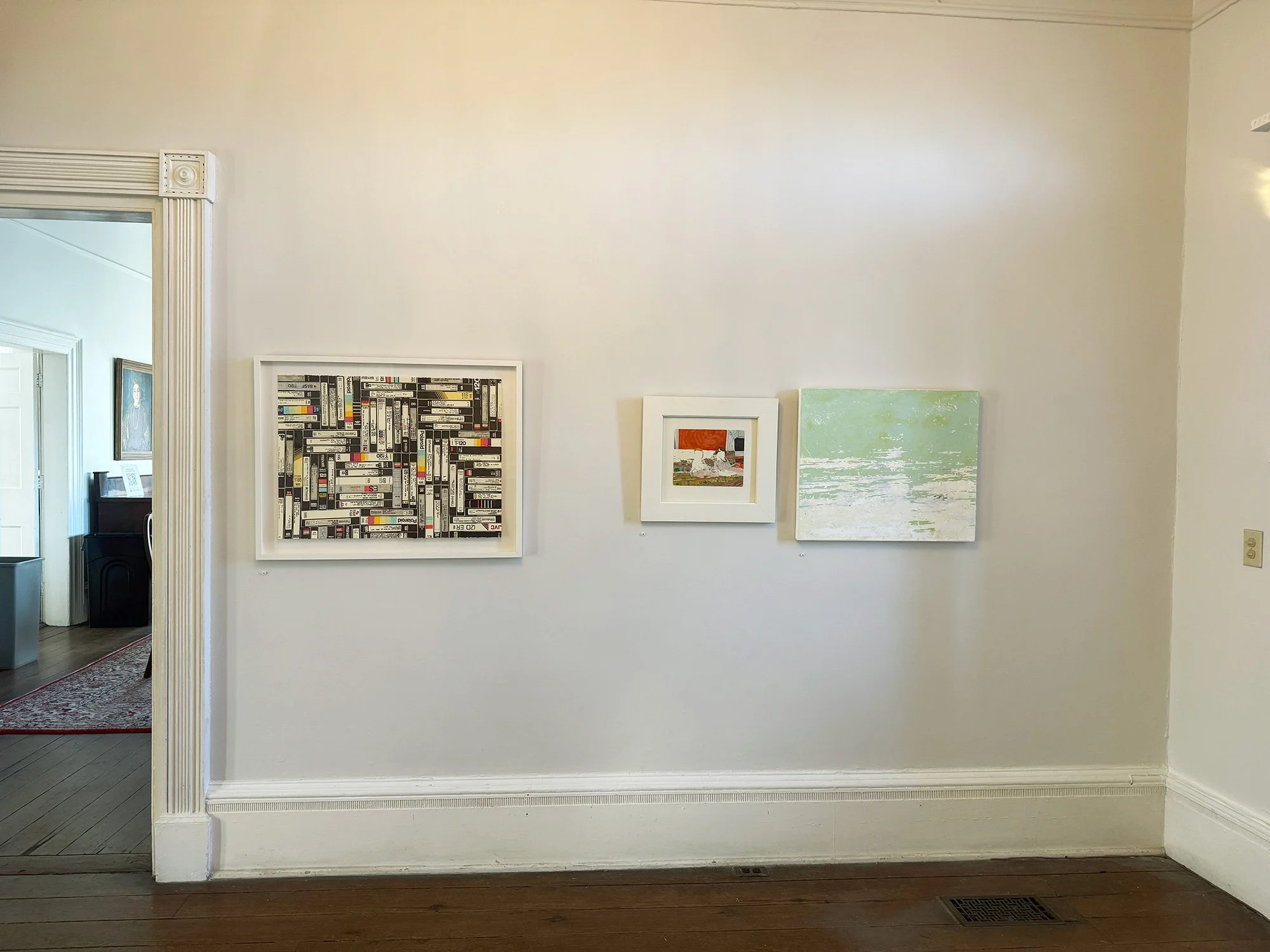 Gallery wall with three framed art pieces in a room with wooden floor and white walls, doorway leading to another room visible on the left.