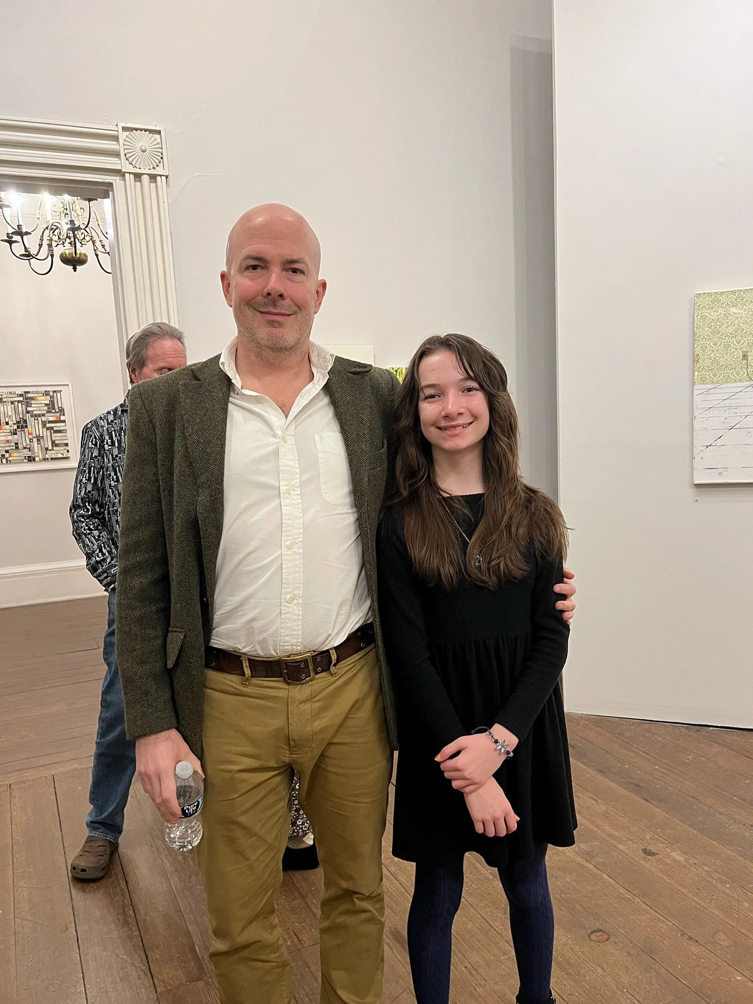 A man and a girl standing side by side in a gallery, smiling at the camera. The man is holding a water bottle and wearing a white shirt with a dark blazer, while the girl is wearing a black dress and tights.