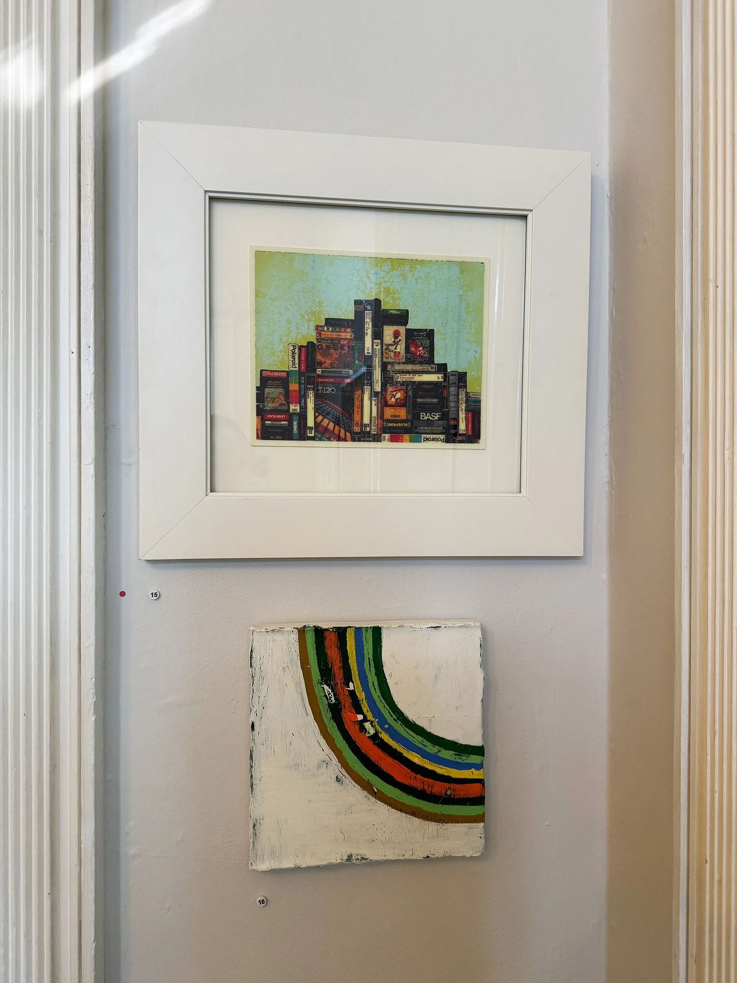 Two pieces of artwork hung on a white gallery wall. The top piece is a framed print of a colorful collage of books and vintage electronics against a green background. The bottom piece is an abstract painting with colorful curved stripes on a white, t