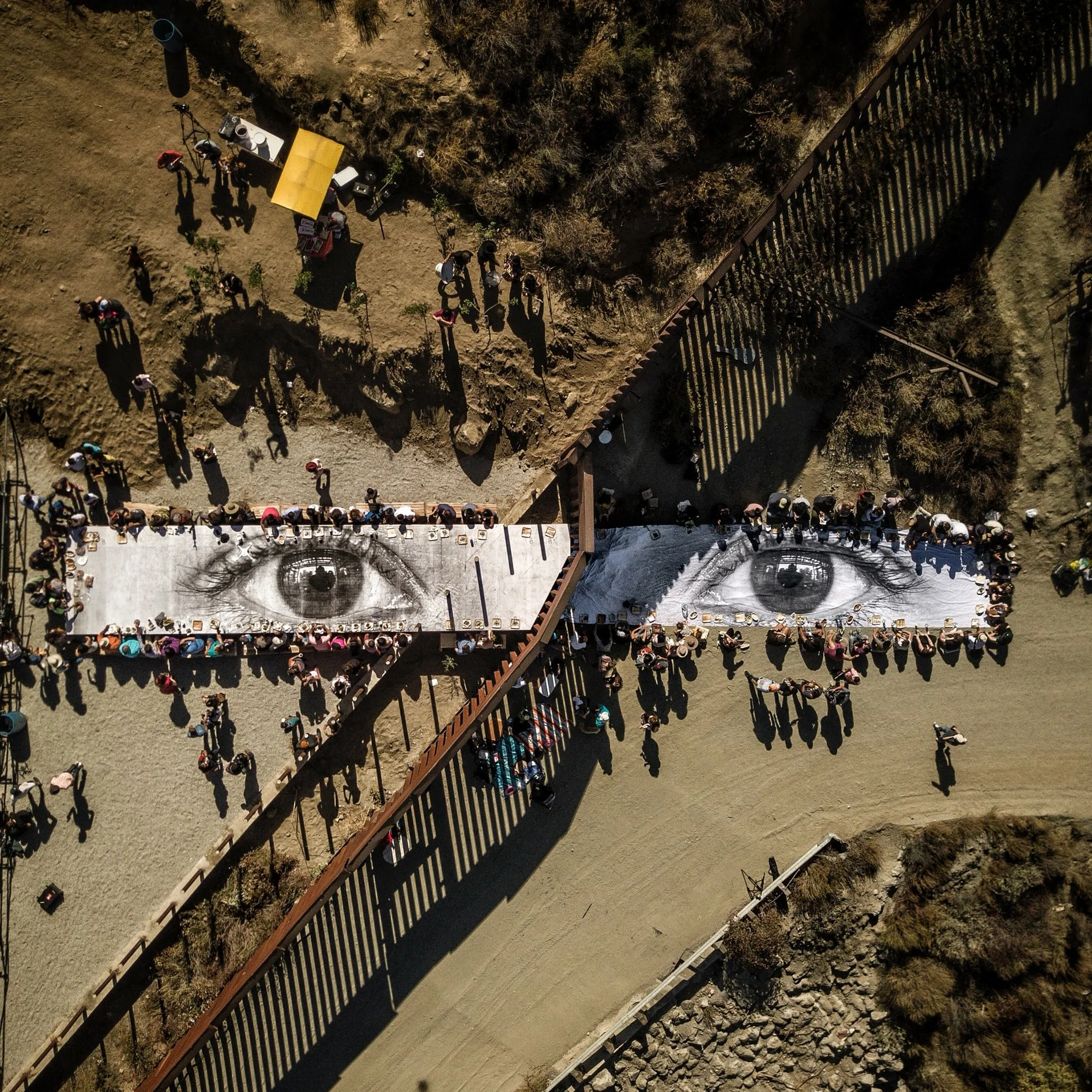 This Overview shows a large picnic at US-Mexico border, spanning from Tecate, California, USA, to Tecate, Baja California, Mexico. The meal is being eaten atop “the eyes of a Dreamer,” a mural painted by French artist “JR” for the one-day-only installation in October 2017. The eyes depicted in the mural are those of a Deferred Action for Childhood Arrivals (DACA) recipient. 32°34'34.1"N, 116°37'45.8"W Source imagery: JR