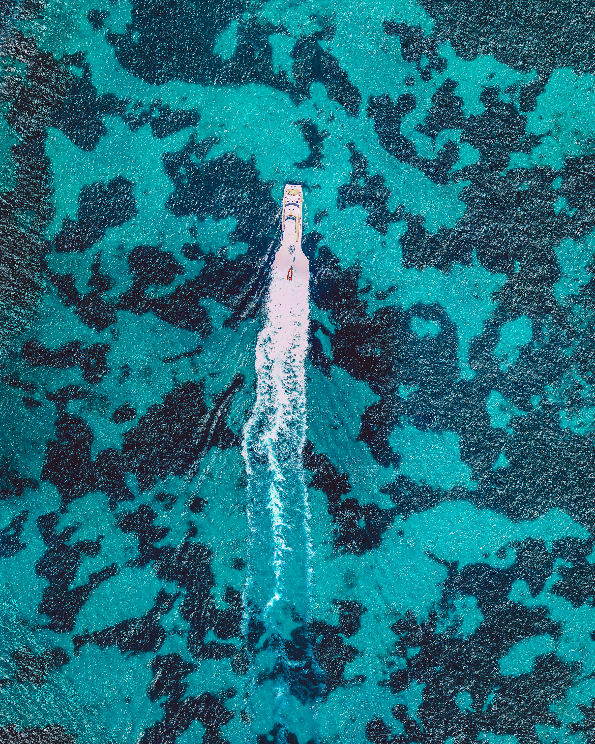 A yacht cruises around Rottnest Island, located off the coast of Perth, Western Australia. The dark blotches seen in the water are parts of the Kingston Reef — one of five Marine Sanctuary Zones surrounding the island. These zones form a habitat for many species of fish and crustaceans, bottlenose dolphins, blue whales, Australian sea lions, New Zealand fur seals, and migrating humpbacks. -31.994995, 115.560582 Source imagery: Nearmap