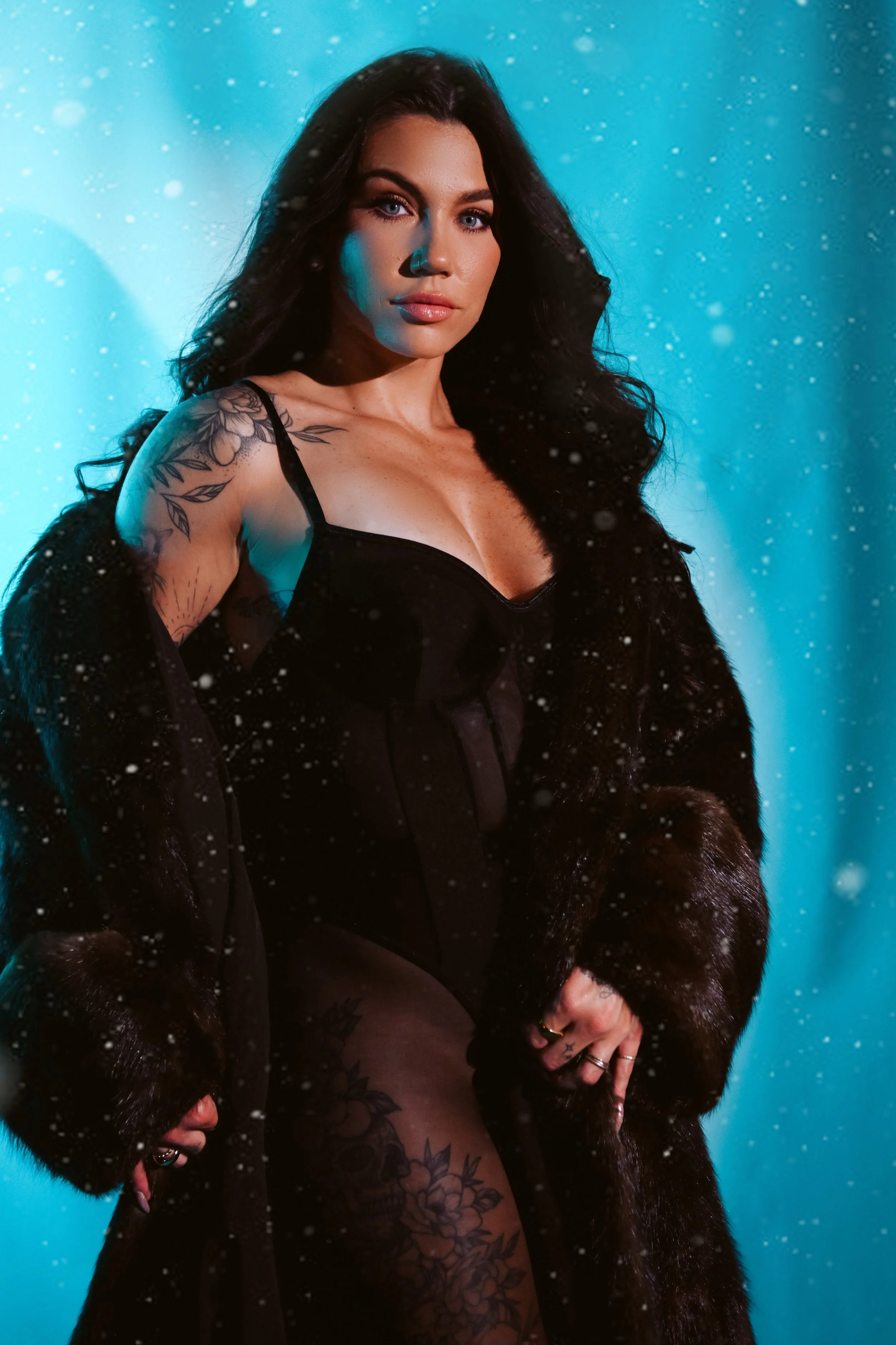 A woman with long dark hair, wearing a black corset, black fur coat, and patterned stockings, posing against a blue background with falling snowflakes.