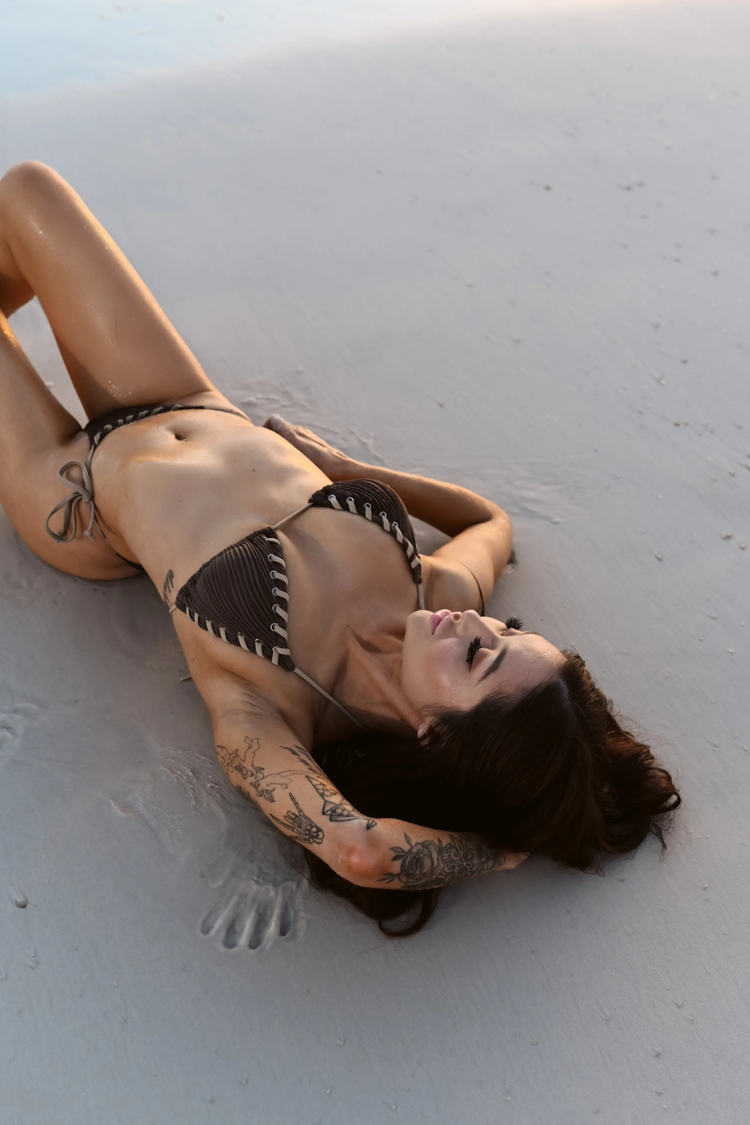 Woman lying on a sandy beach in a black bikini with tattoos, relaxing with her eyes closed.