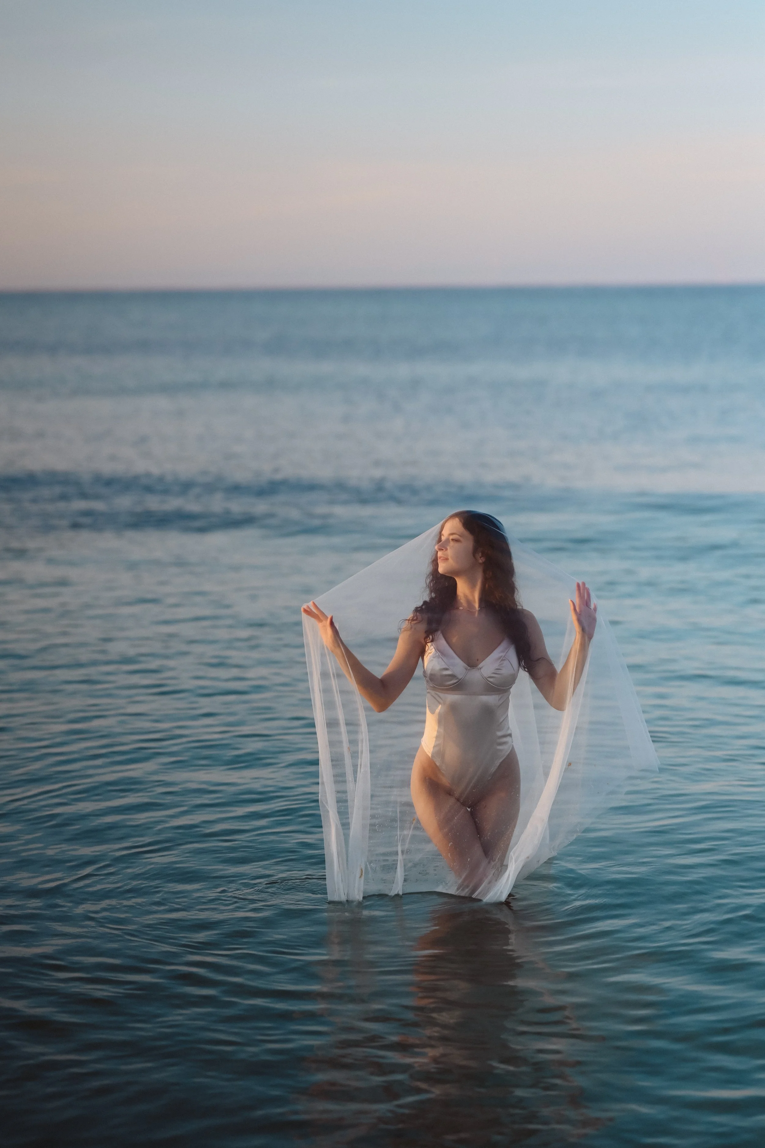 A woman in a white dress standing in ocean water, holding a veil over her head during sunset.