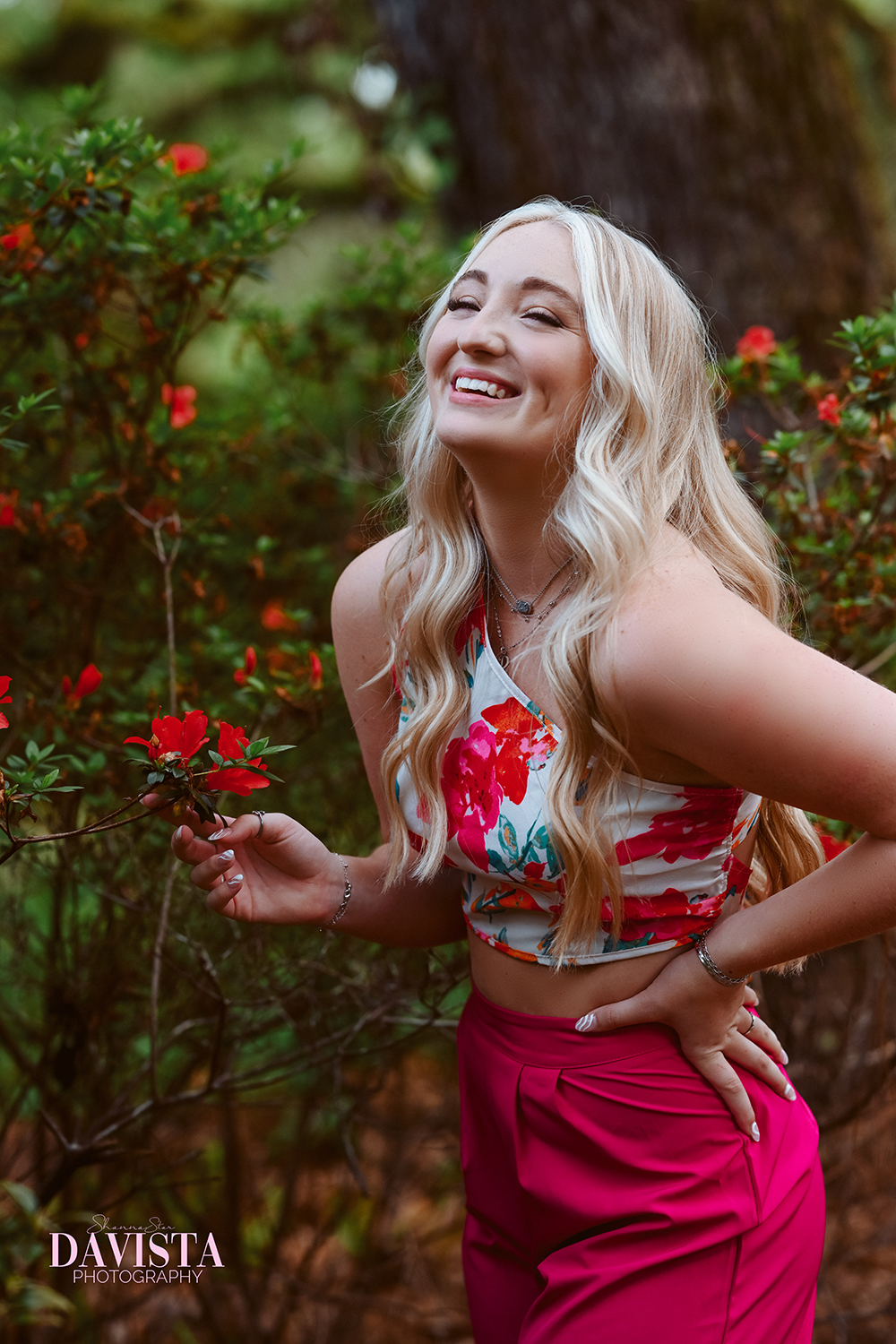 A young woman with long blonde hair in loose waves, smiling joyfully with her eyes closed, outdoors in a garden setting with green foliage and red flowers, wearing a floral crop top and bright pink pants.
