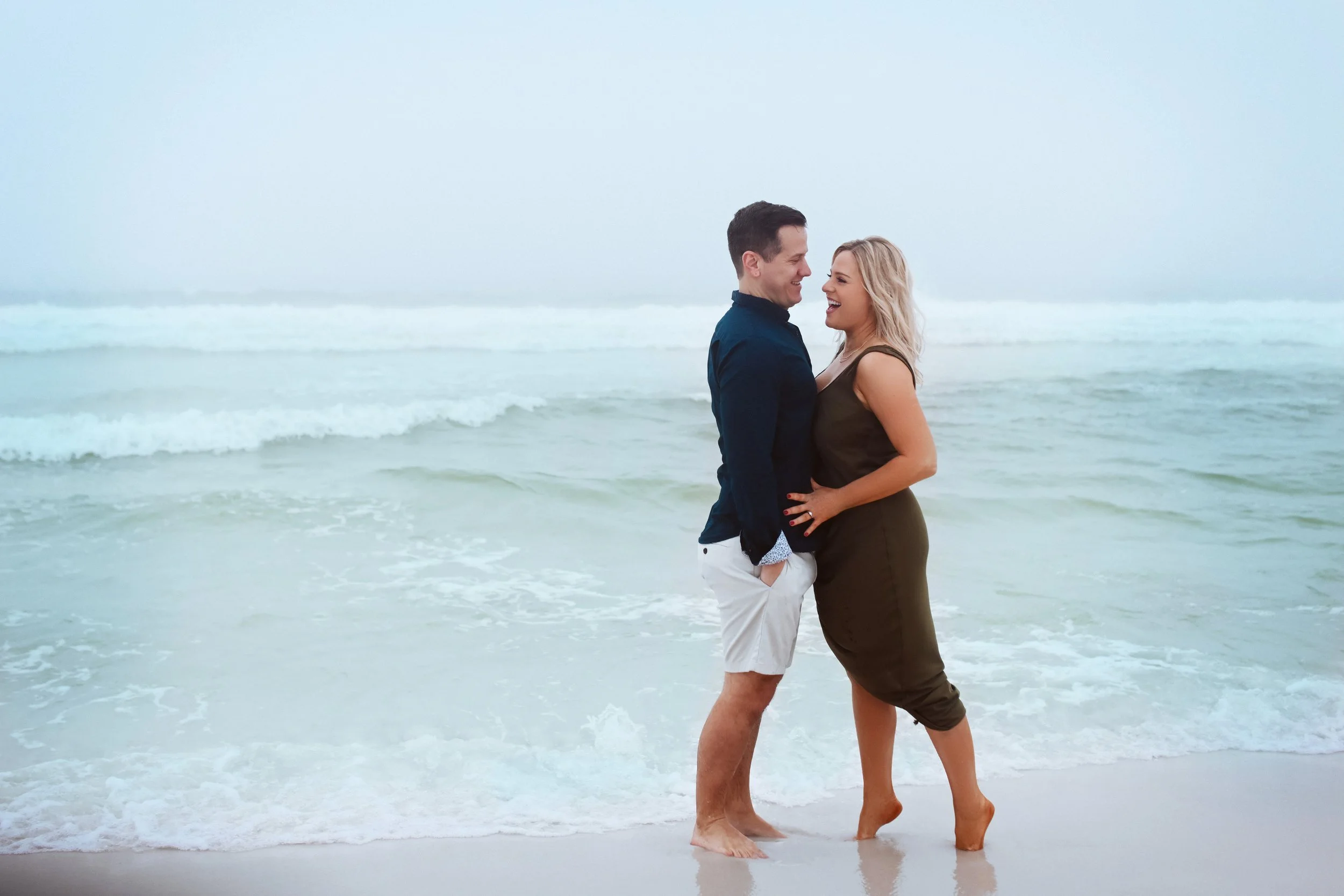A couple standing barefoot on the beach, smiling and looking at each other with the ocean and waves in the background.