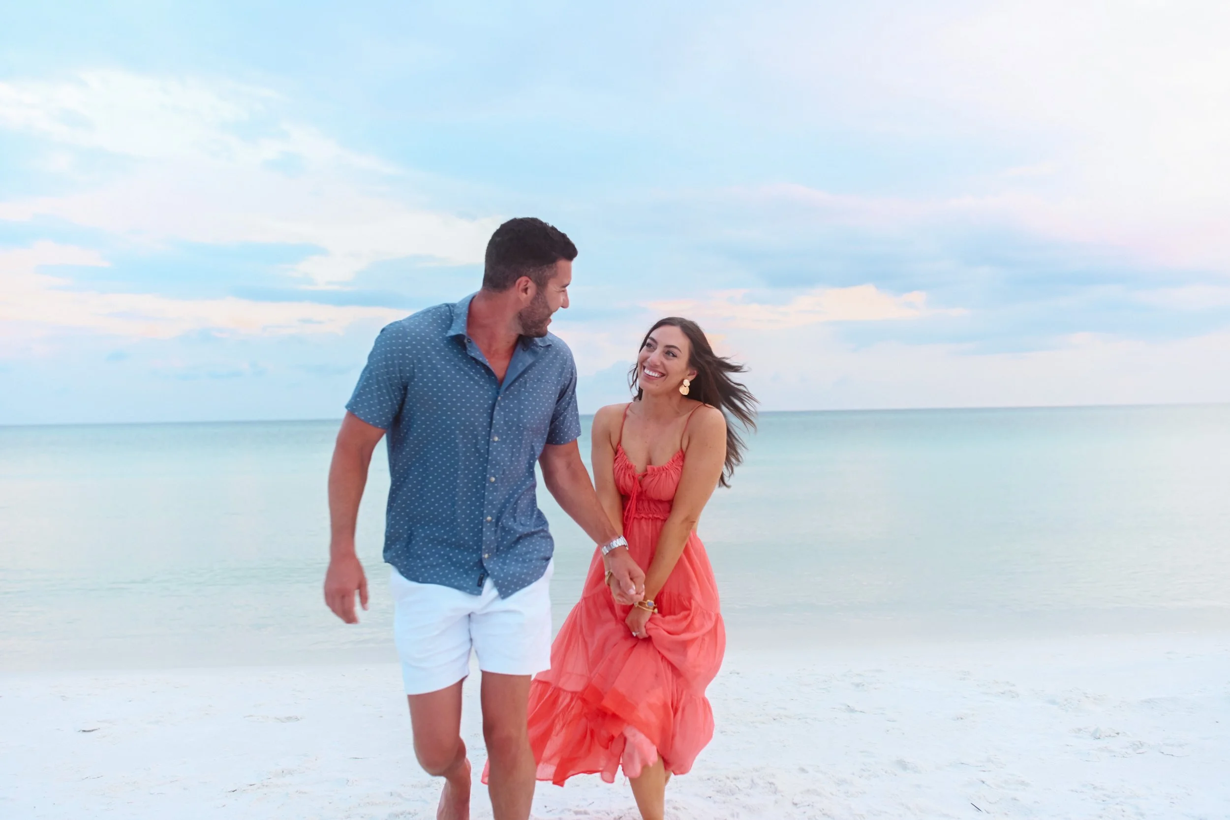 A smiling couple walking together on the beach, the man holding the woman's hand, with the ocean and a partly cloudy sky in the background.